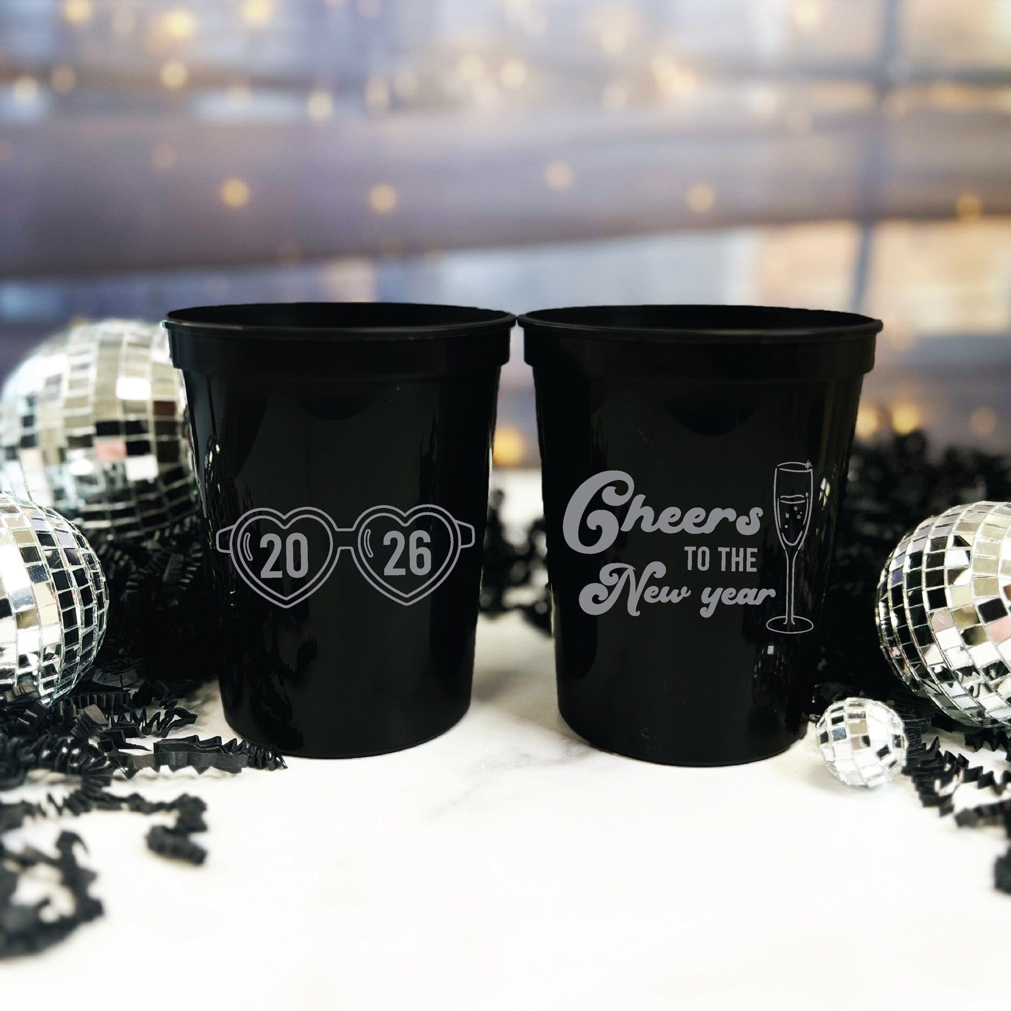 Two black plastic stadium cups are personalized with festive designs, one featuring stylized heart-shaped glasses and the other a champagne flute graphic, set against a backdrop of disco balls and decorative crinkle paper for a party.