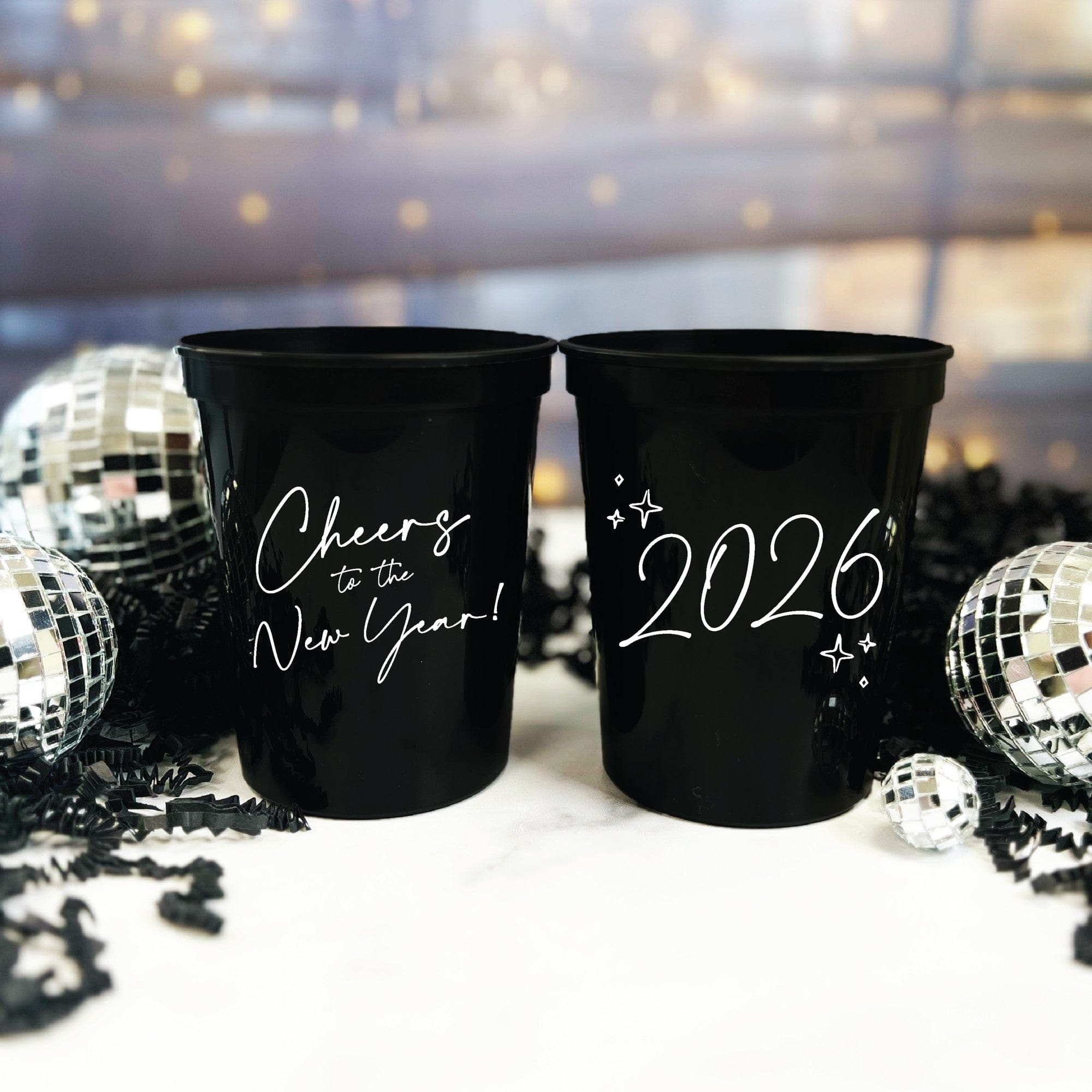 Two black personalized plastic party cups for a New Year's celebration, decorated with white script and displayed amidst disco balls and shredded paper with a festive bokeh background.
