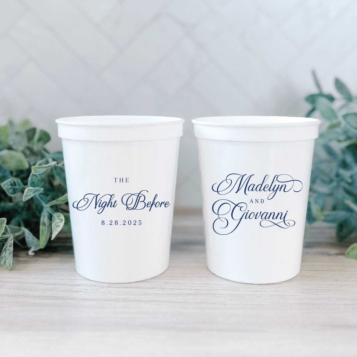 Two white stadium cups with elegant script text reading "The Night Before" and date on one cup, "Madelyn and Gerannt" on the other, displayed with greenery.