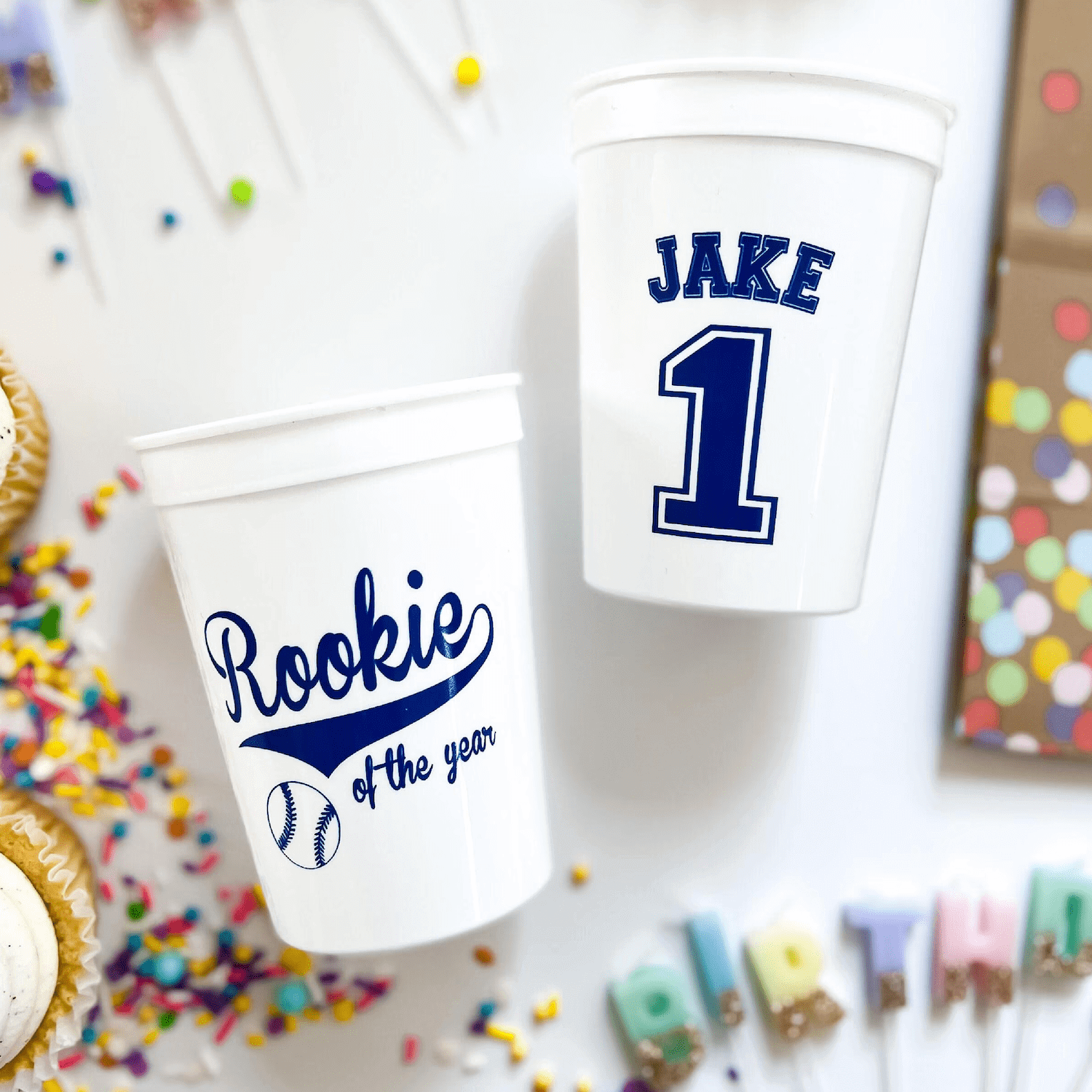Two white stadium cups personalized with "Rookie of the Year" and baseball design on one, "Jake 1" on the other, surrounded by colorful confetti and party decorations.