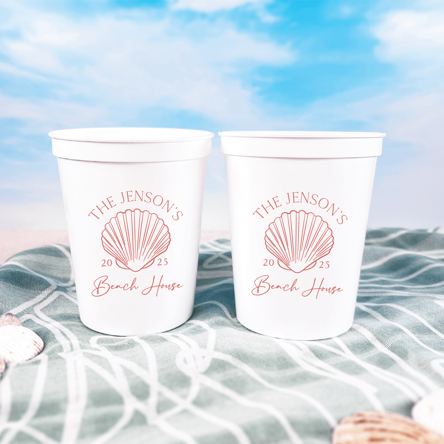 Two white stadium cups with pink seashell design and "The Jensons Beach House" personalized text, displayed on beach netting outdoors.