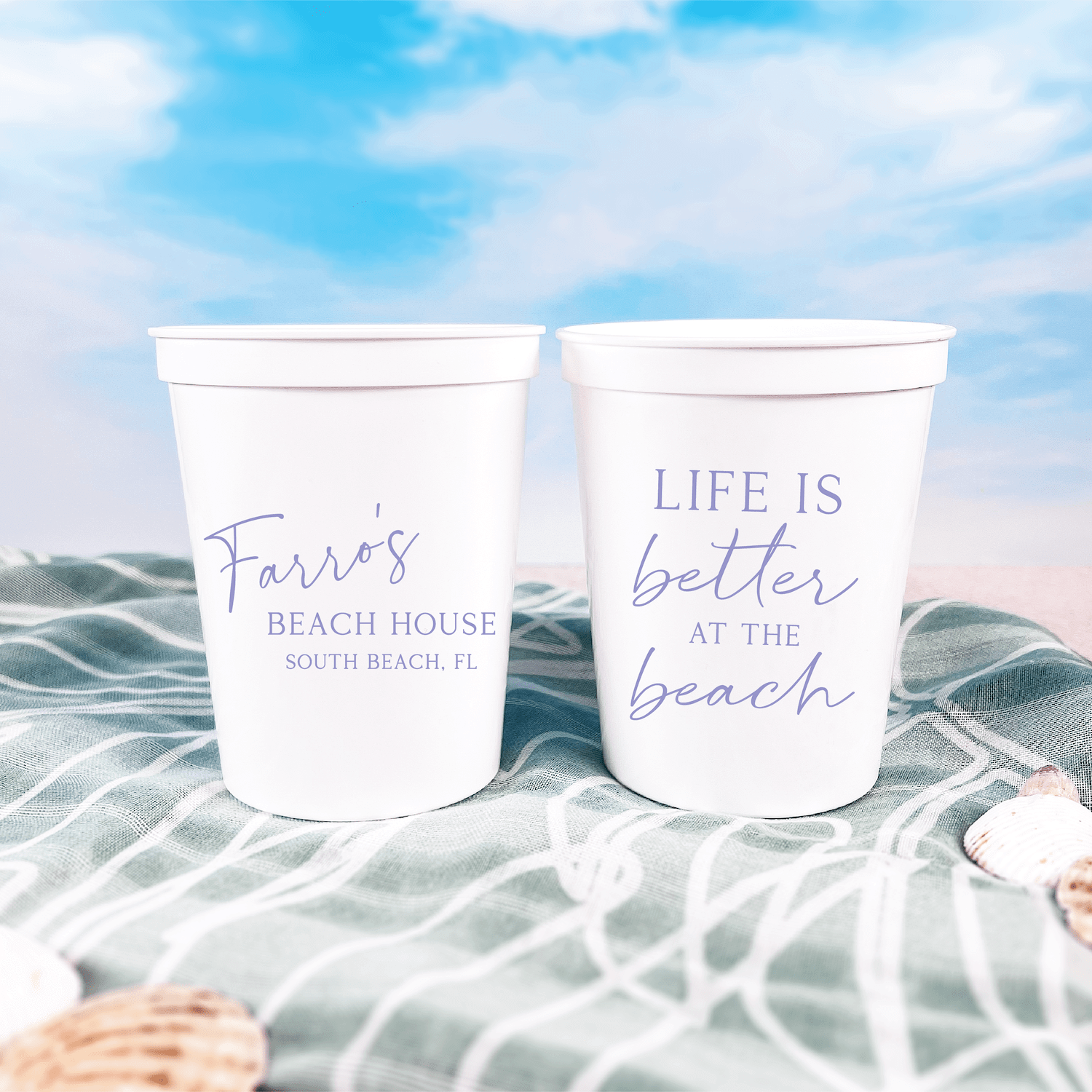 Two white stadium cups on a beach blanket with personalized text reading "Favors Beach House South Beach, FL" and "Life is Better at the Beach" in purple lettering