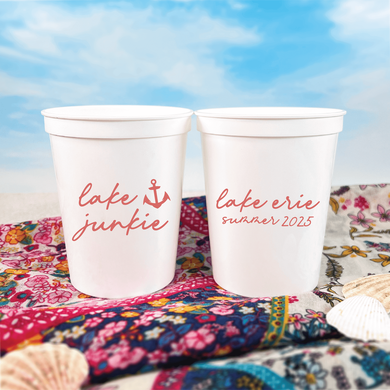 Two white stadium cups with red script text reading "cafe junkie" and "cafe eerie summer 2025" displayed on colorful patterned fabric outdoors.