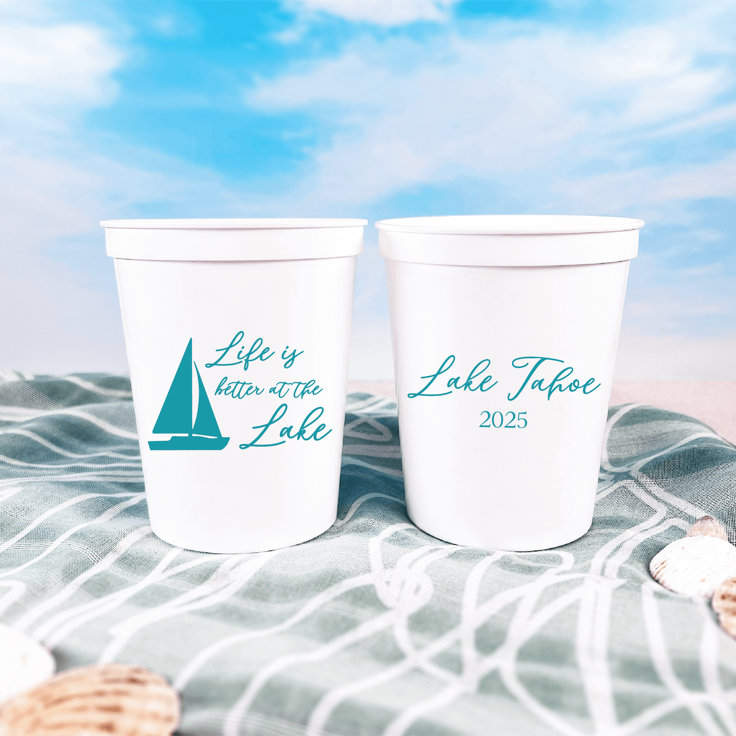 White stadium cups with teal sailboat and "Life is Better at the Lake" text on left cup, "Lake Tahoe 2025" on right cup, displayed on beach towel.