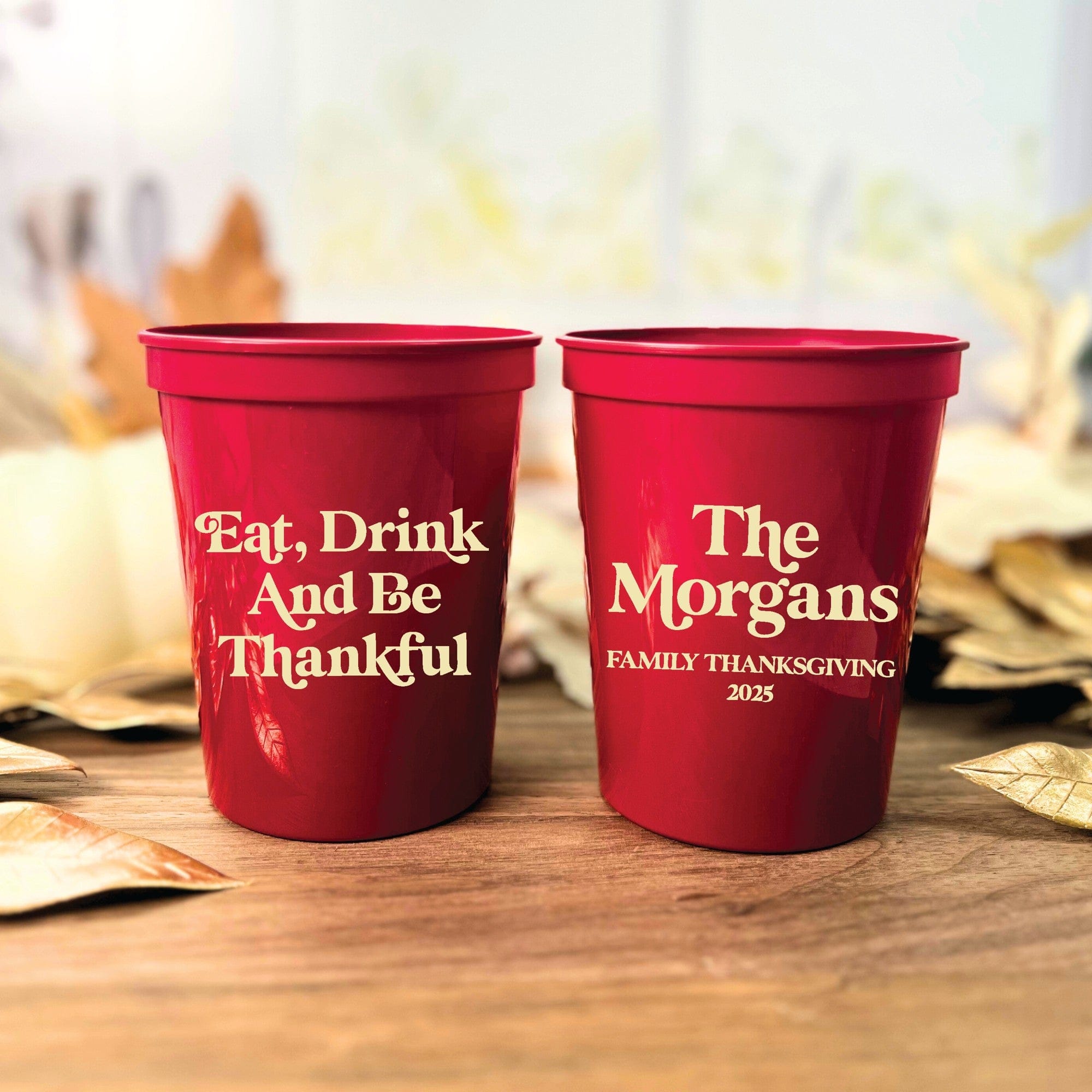Two personalized red plastic party cups are displayed on a wooden surface surrounded by autumn leaves, ideal for a Thanksgiving celebration.