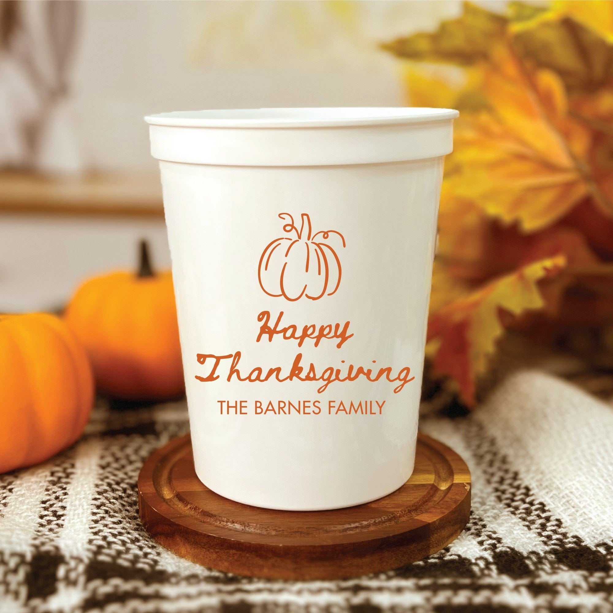 A white plastic stadium cup featuring an orange pumpkin design for a Thanksgiving celebration is displayed on a wooden coaster, surrounded by small decorative pumpkins and fall foliage.