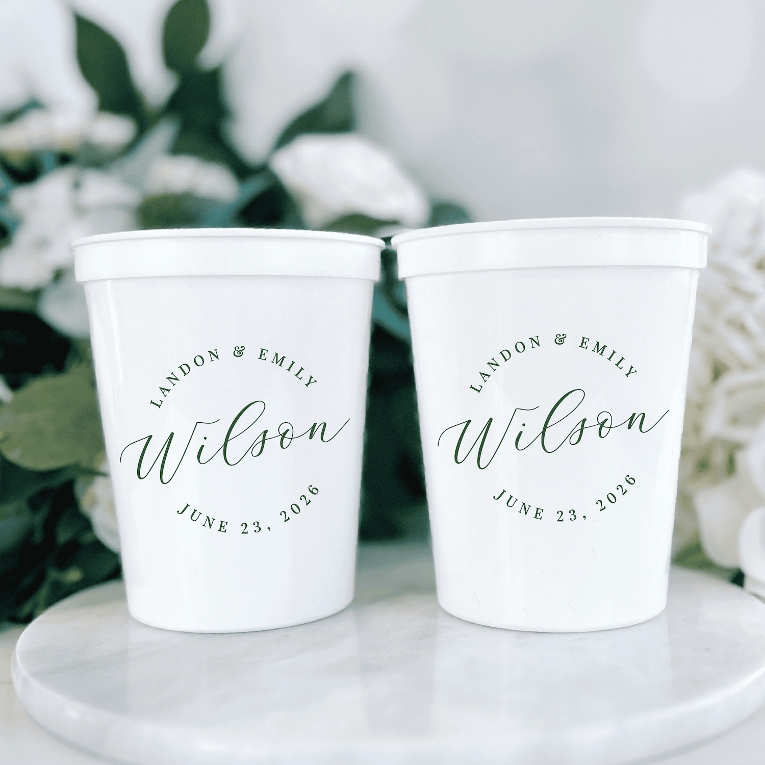 White stadium cups personalized with "Wilson" and wedding date June 23, 2024, displayed on marble surface with greenery.