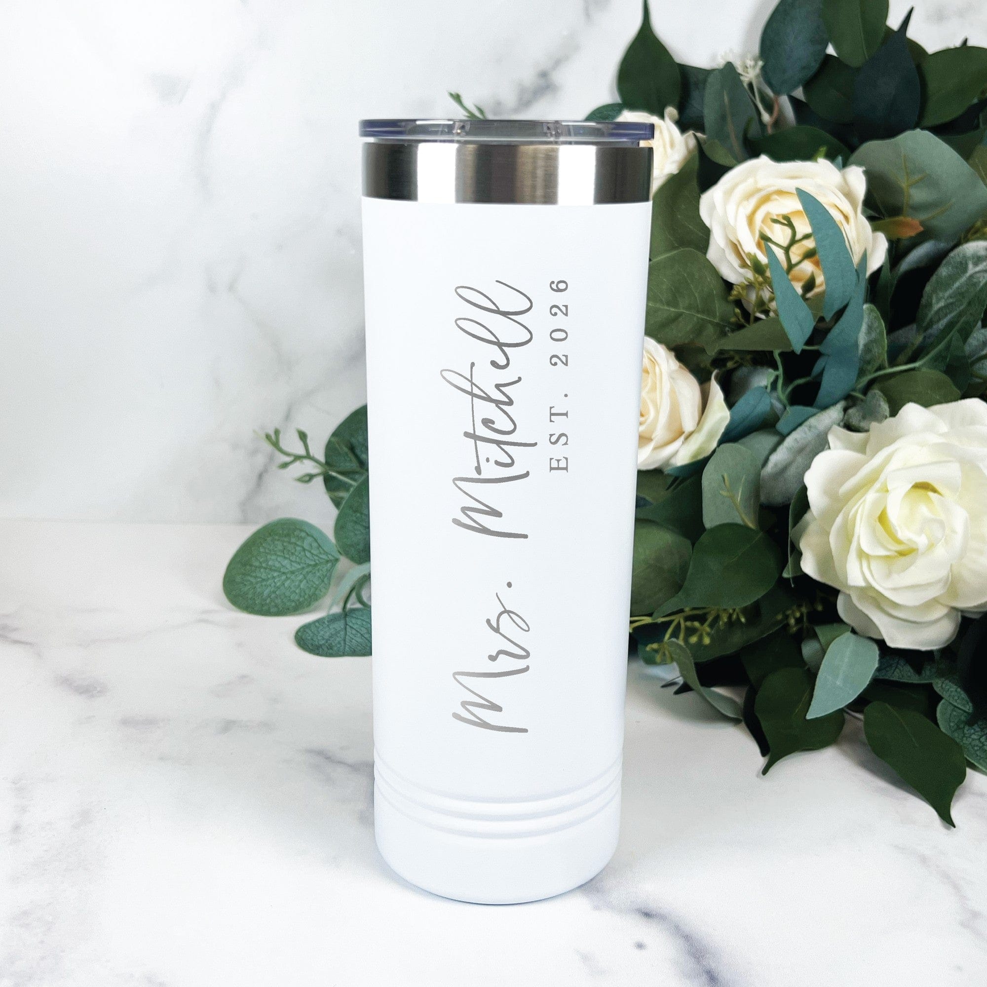 A tall, white insulated stainless steel tumbler with a metallic rim and clear lid, personalized with elegant script text for a bridal event.