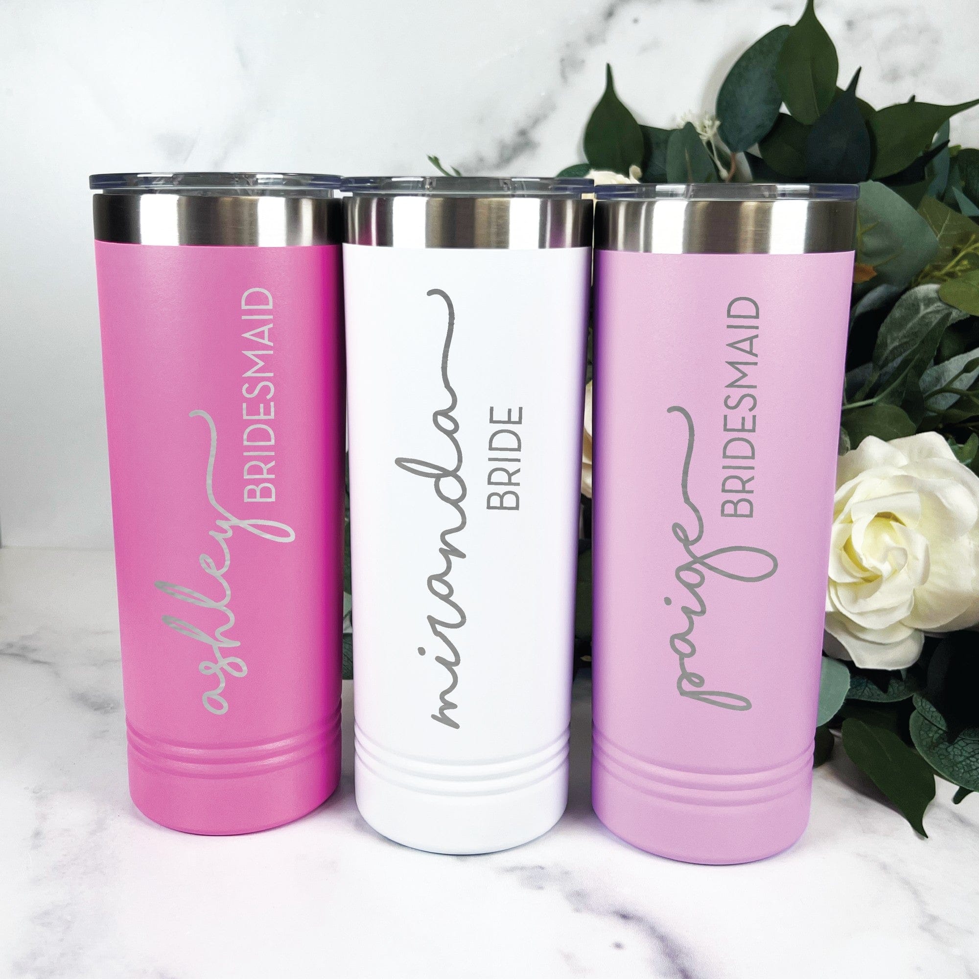 Three personalized skinny stainless steel tumblers, two in shades of pink and one in white, each with a silver rim and clear lid, ideal for wedding party gifts.