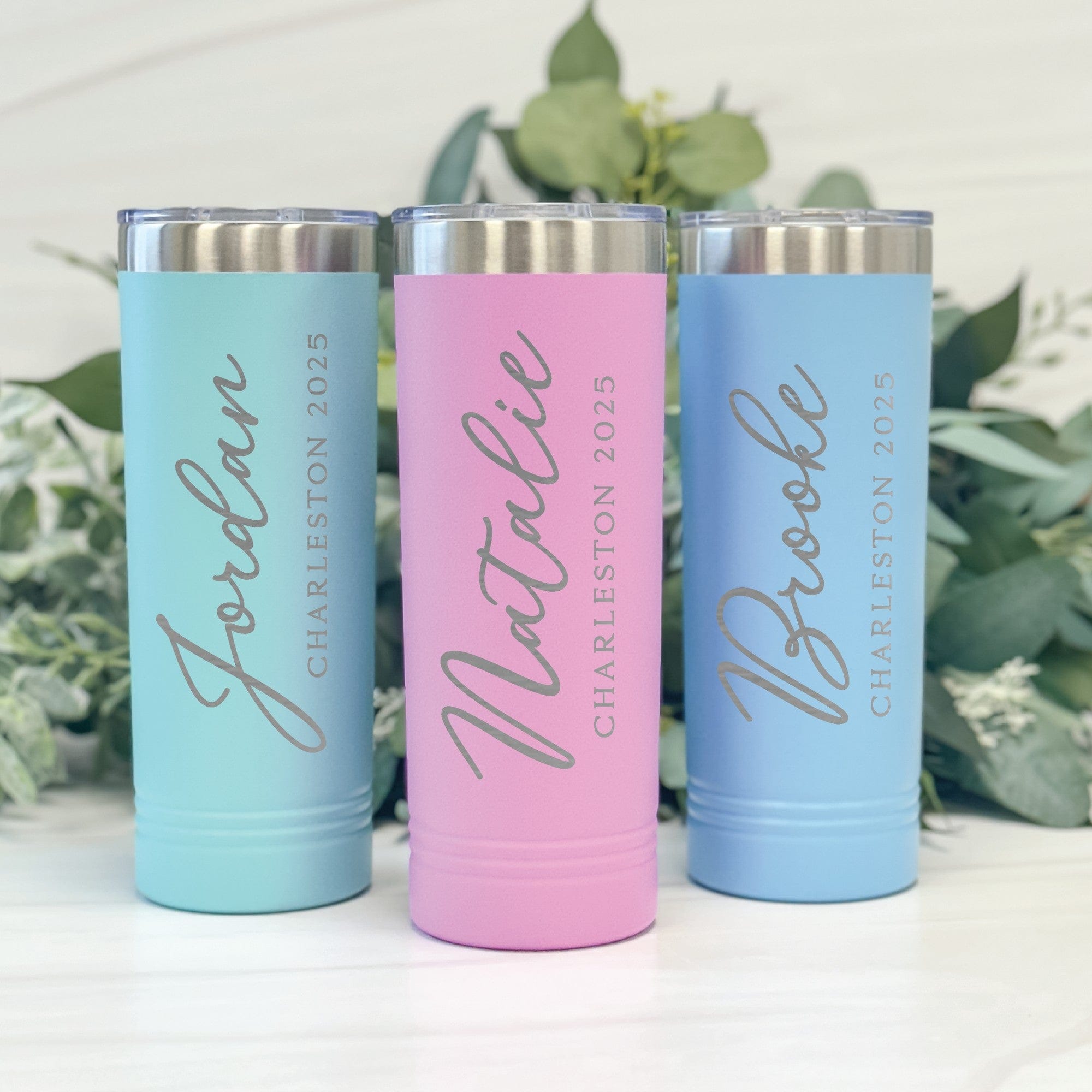 Three tall, slender, insulated tumblers in pastel aqua, pink, and light blue, crafted from stainless steel with clear, spill-resistant lids, designed as personalized drinkware for wedding parties or special events.