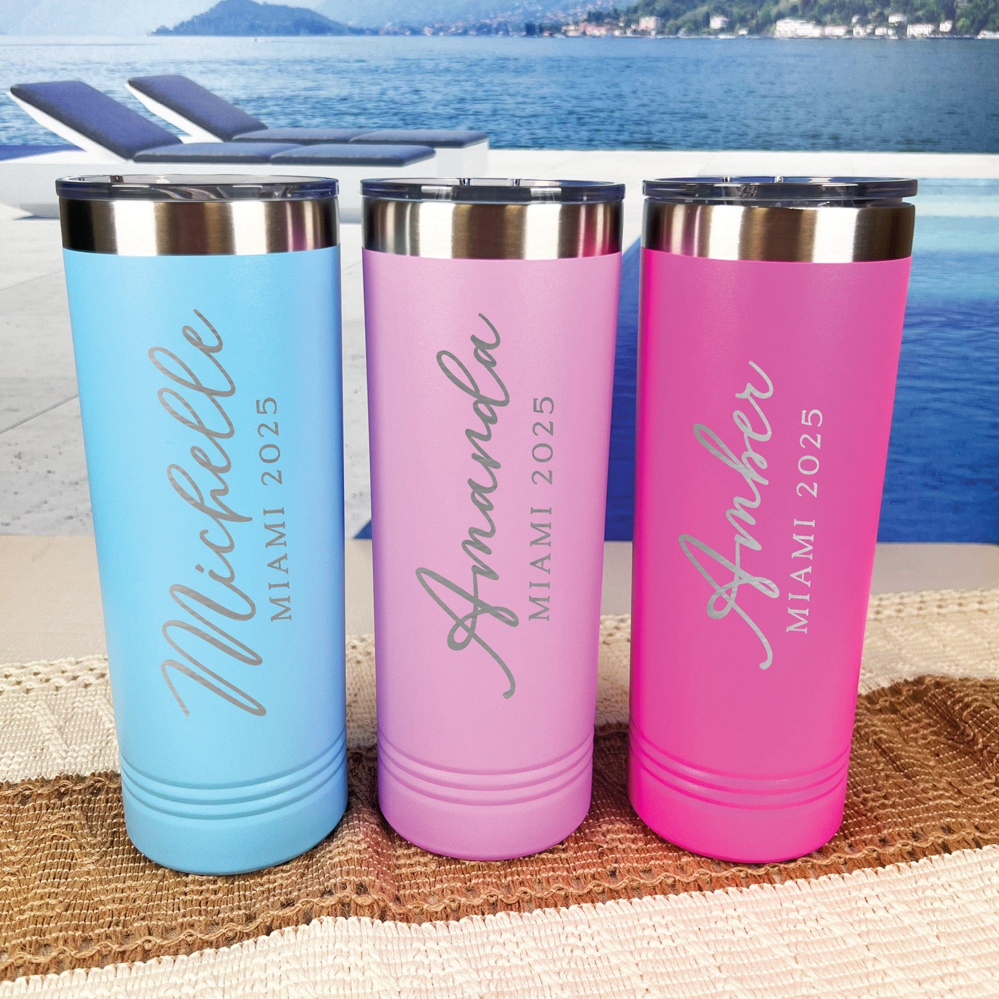 Three vibrantly colored insulated tumblers, including light blue, light pink, and fuchsia, each featuring custom engraved text and topped with a stainless steel lid, displayed on a textured mat with a tropical water background, ideal for a destination wedding or bachelorette party.