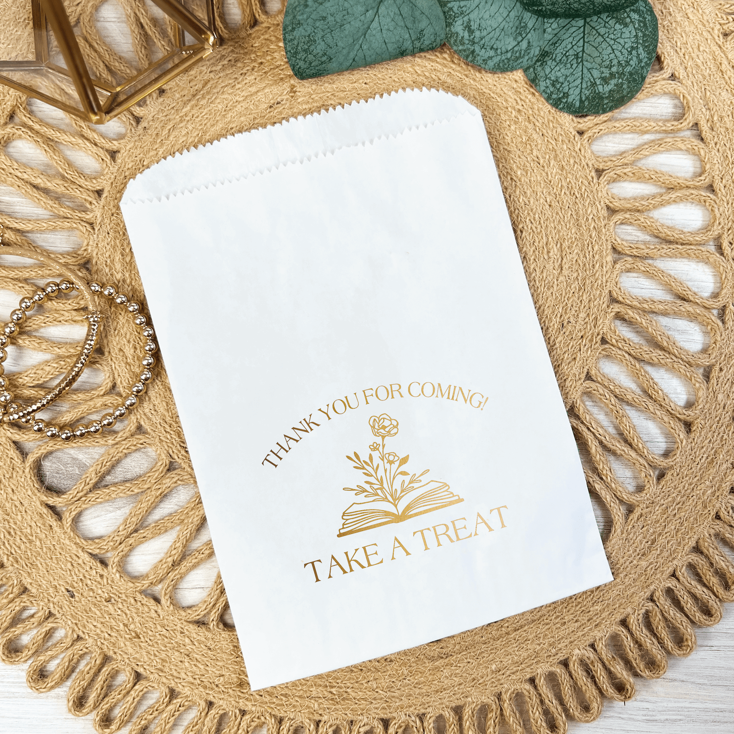 White paper treat bag with gold foil-stamped text reading "THANK YOU FOR COMING" and "TAKE A TREAT" with floral design, displayed on woven placemat.