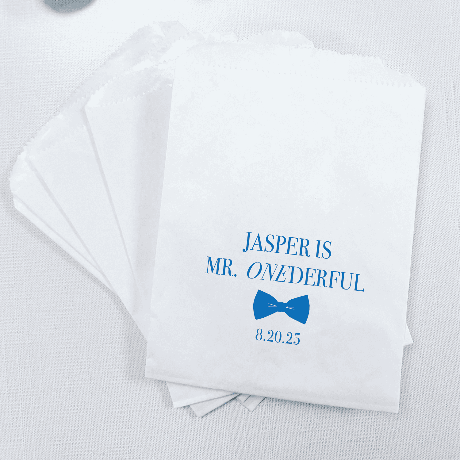 White treat bags with blue text reading "Jasper is Mr. Onederful" and a blue bow tie graphic with date 8.20.25