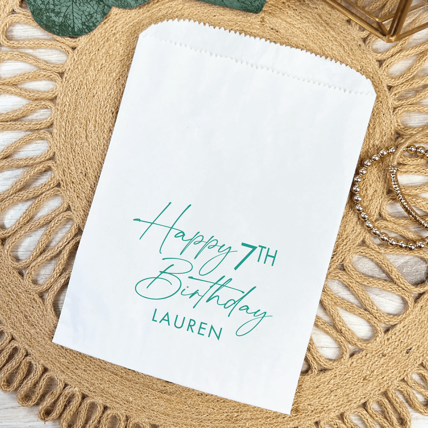 White paper treat bag with teal handwritten text reading "Happy 7TH Birthday" and "LAUREN" on natural woven placemat.