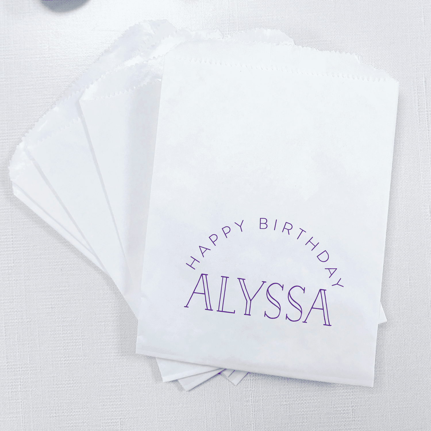 Stack of white paper treat bags with purple text reading "Happy Birthday Alyssa"