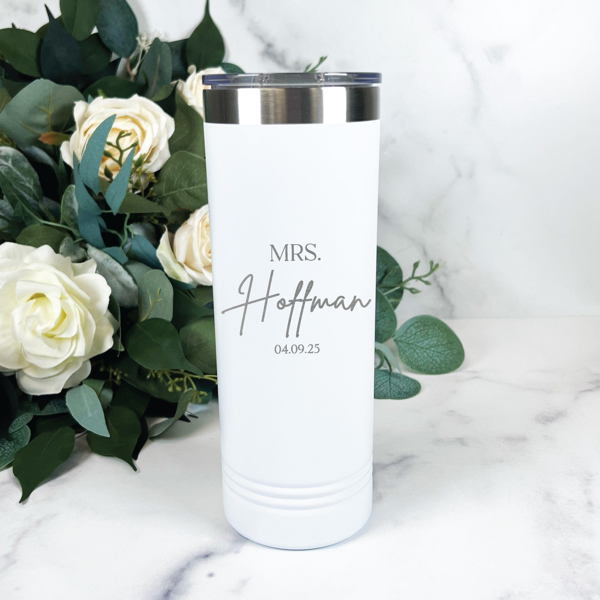 A white personalized insulated tumbler with a silver rim and clear lid, featuring elegant lettering, displayed on a marble surface with white roses and eucalyptus, perfect for a bridal event.