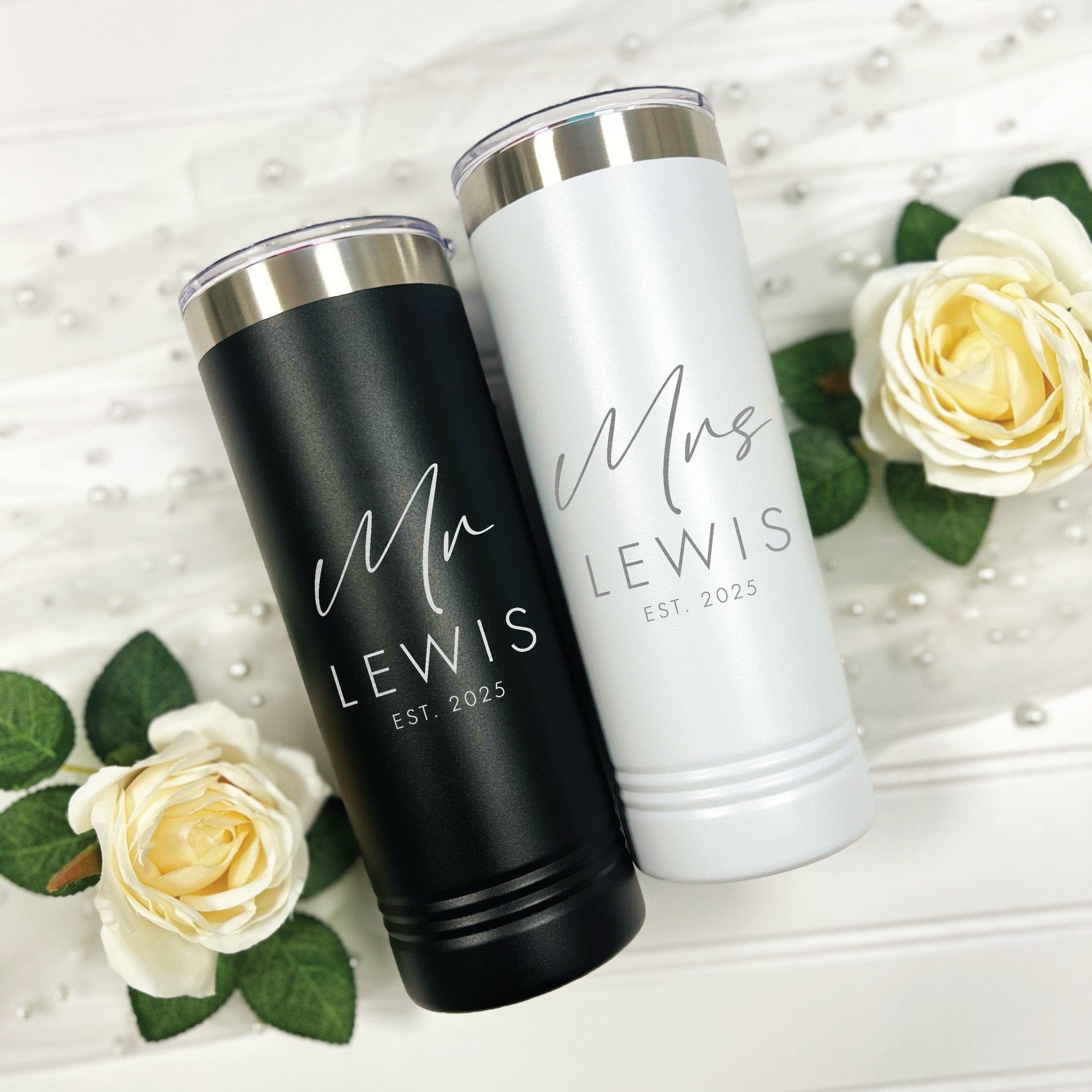 A matching set of insulated metal skinny tumblers, one matte black and one matte white with silver rims and clear lids, ideal for a wedding or bridal celebration.