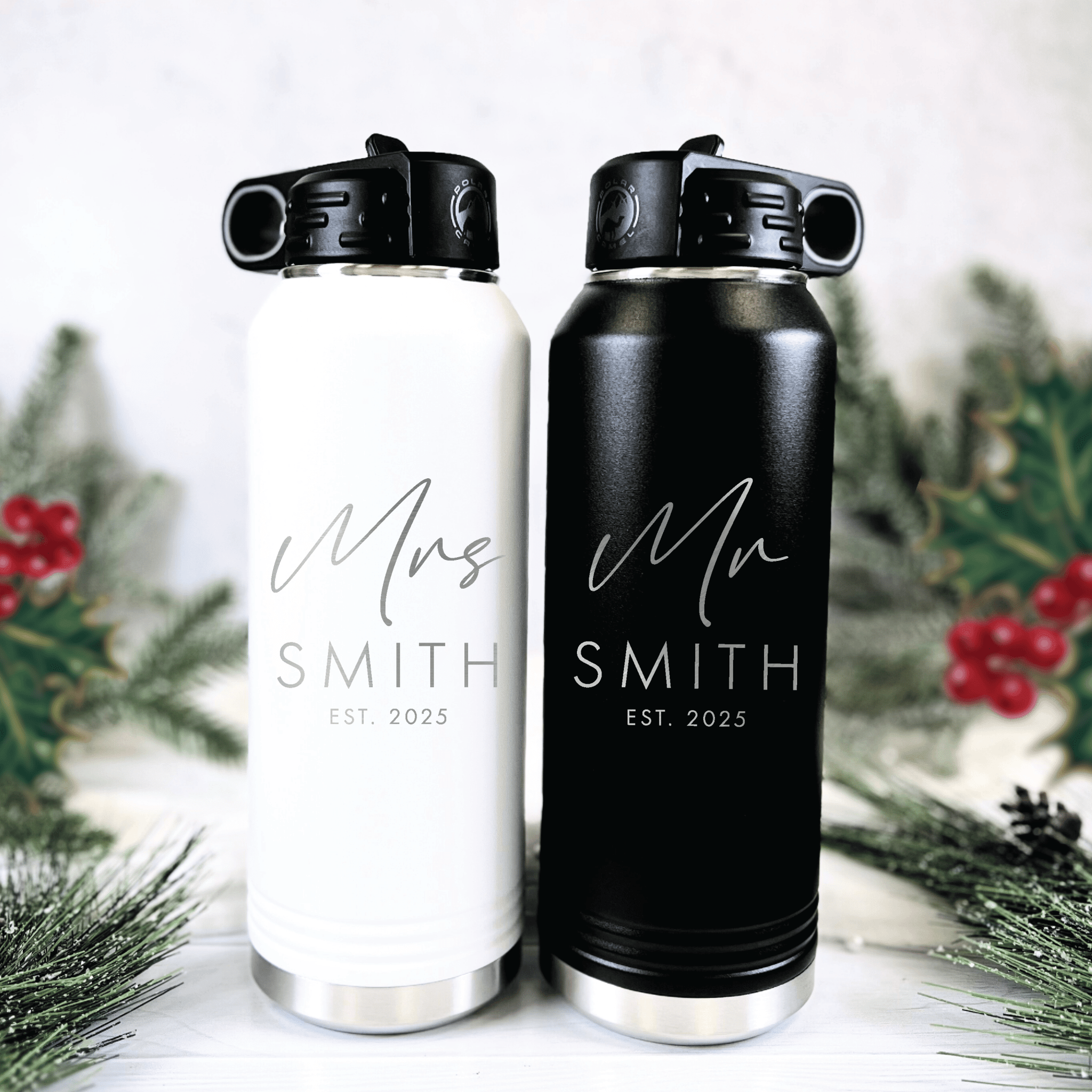White and black insulated water bottles with "Mrs Smith EST 2025" and "Mr Smith EST 2025" engraved, displayed with evergreen sprigs and red berries.
