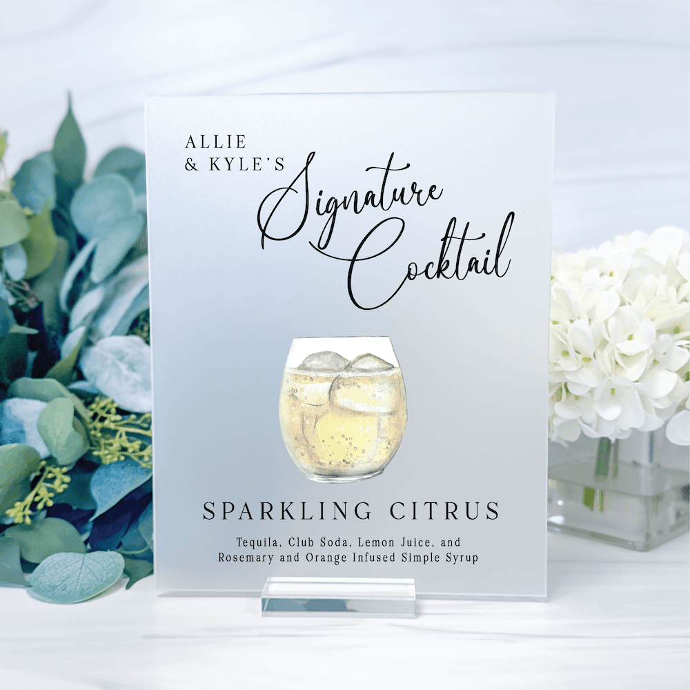White card displaying "Allie & Kylie's Signature Cocktail" with sparkling citrus drink illustration and ingredients list, surrounded by greenery and white flowers.