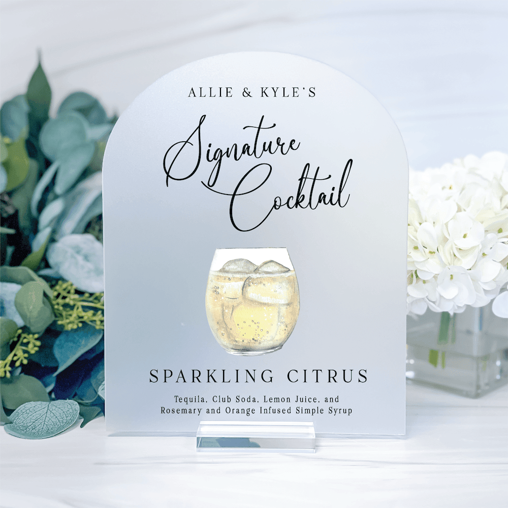 White arched cocktail menu card with "Allie & Kyle's Signature Cocktail" text and sparkling citrus drink illustration, flanked by greenery and white flowers.