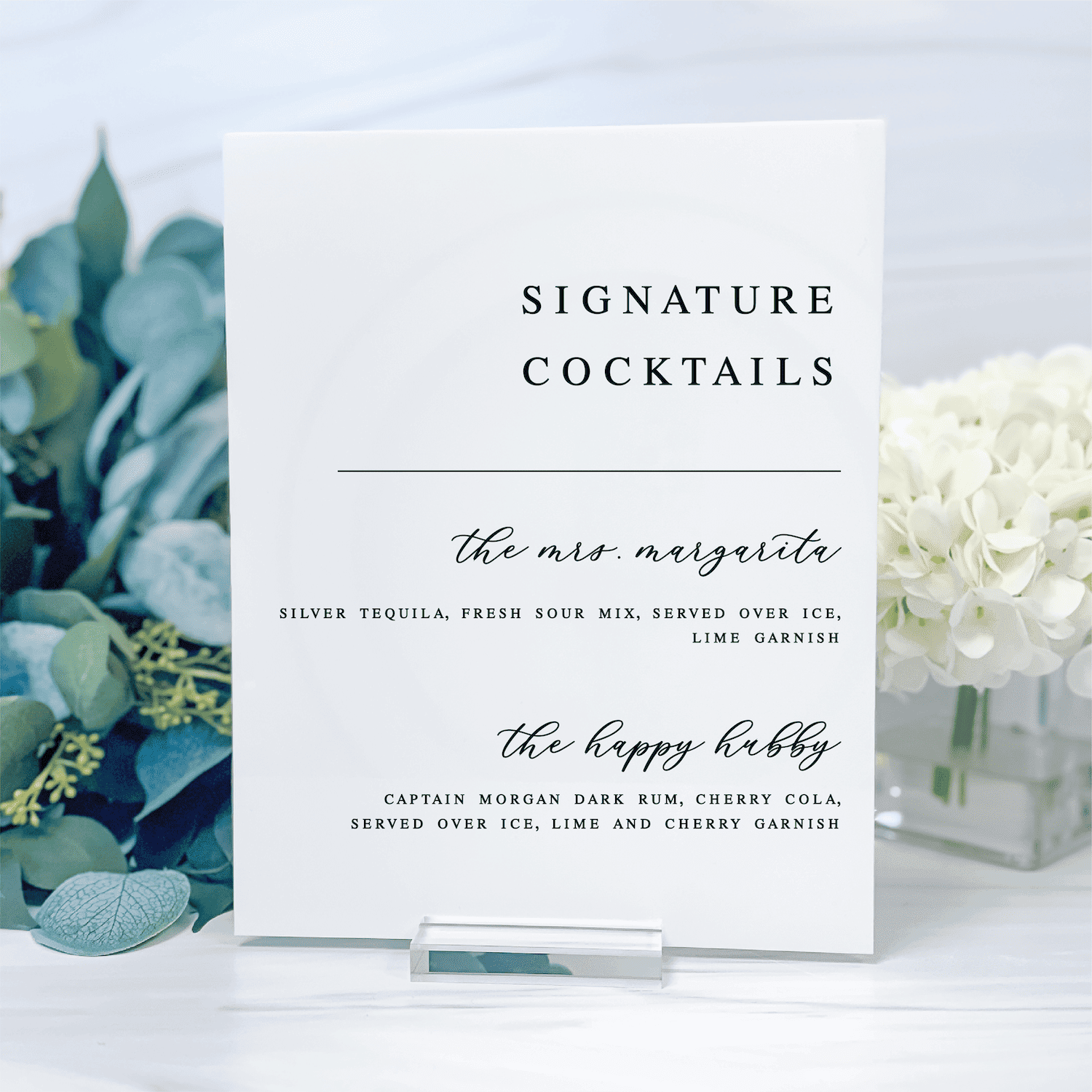 White acrylic bar sign displaying "Signature Cocktails" with two drink recipes, flanked by greenery and white flowers.