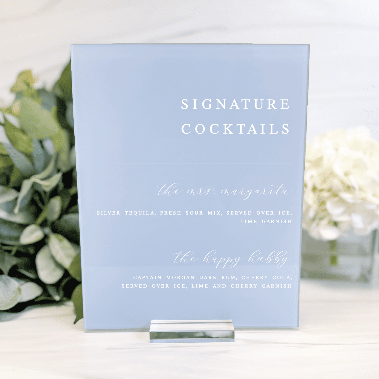 Pale blue acrylic bar sign with white text reading "Signature Cocktails" and two drink recipes below, displayed on a clear stand with greenery and white flowers.