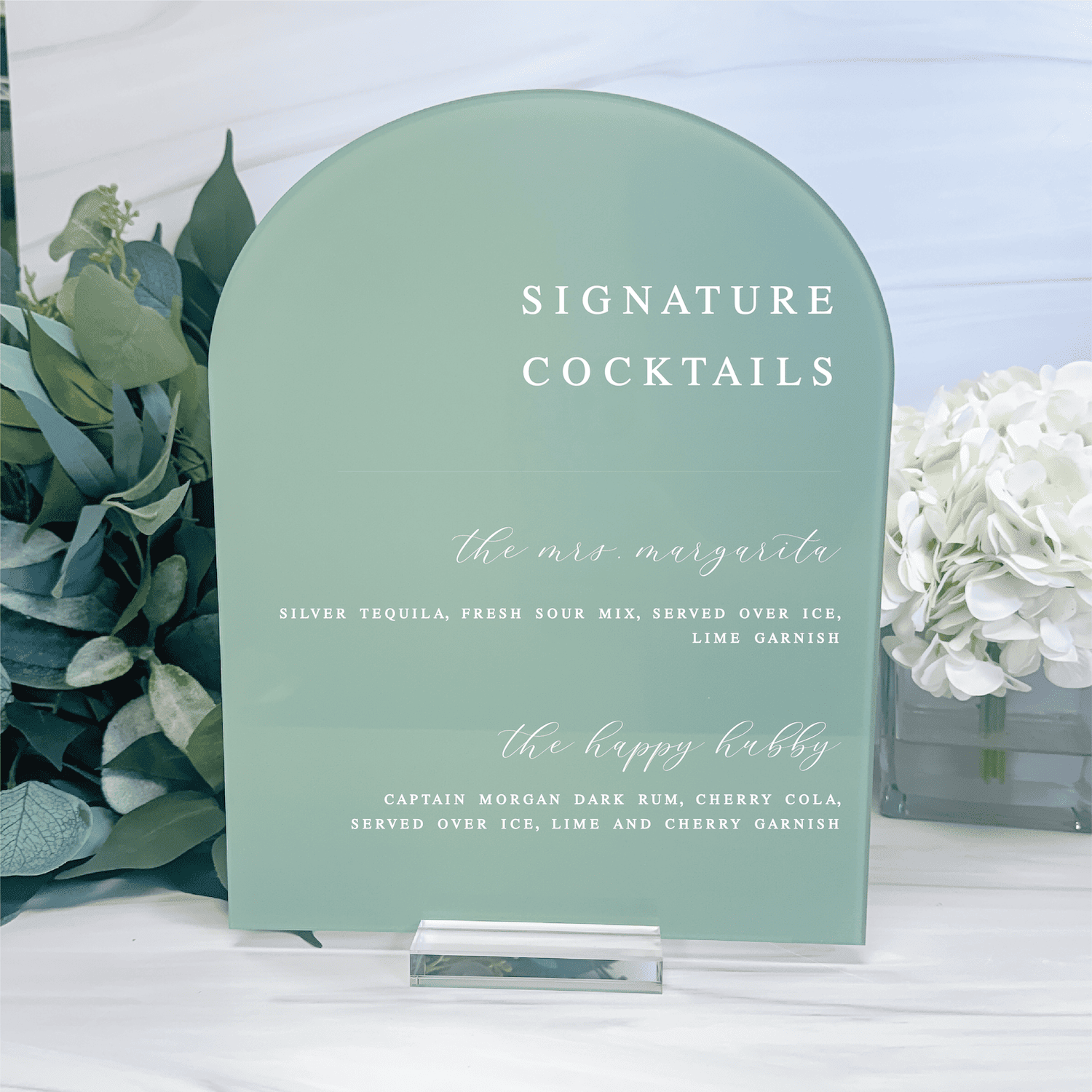 Sage green arched acrylic bar sign with "Signature Cocktails" header and two drink recipes in white script and serif text, displayed with greenery and white flowers.