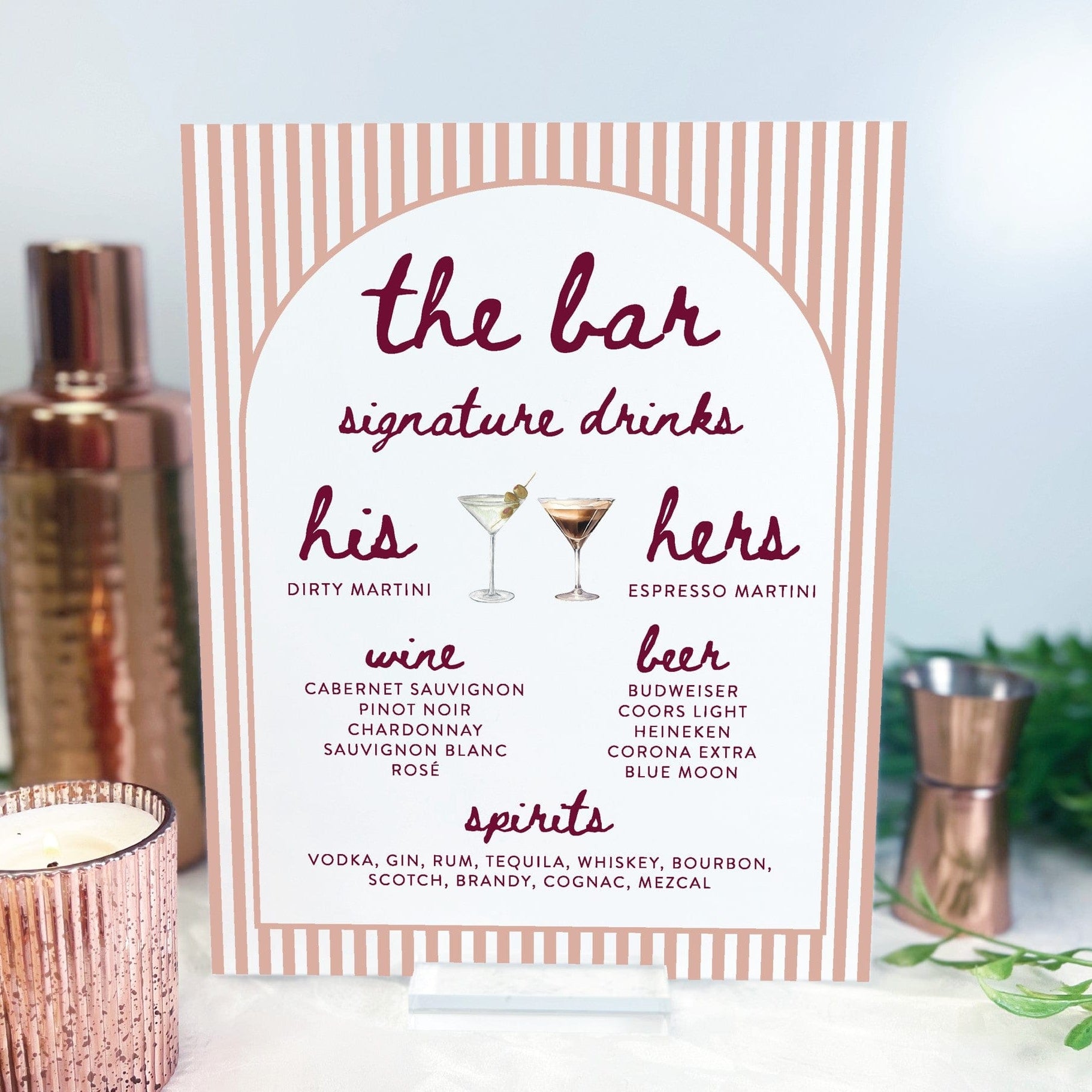 A blush and white striped bar menu sign with dark red lettering, displaying various drink options for a wedding or event, presented on a clear acrylic stand.