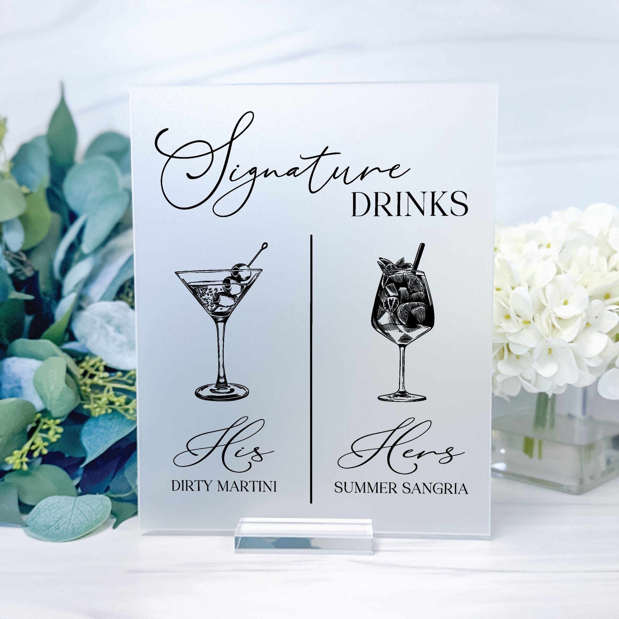 A rectangular frosted acrylic signature drinks sign featuring two illustrated cocktails and text, displayed on a clear acrylic stand, suitable for a wedding or event bar.