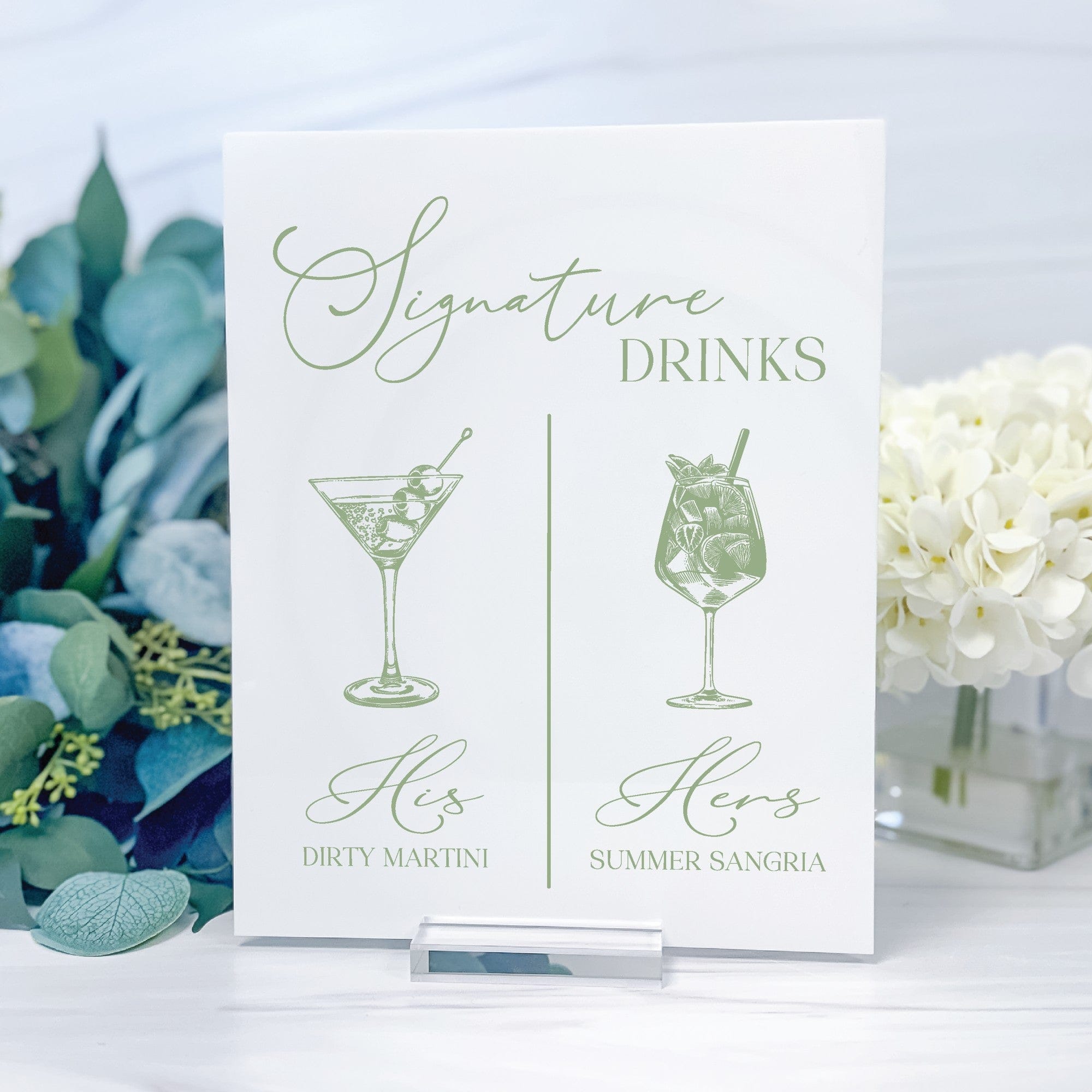 A personalized white signature drinks sign featuring green calligraphy and illustrations of two cocktails, presented in an acrylic stand for a wedding or event bar.
