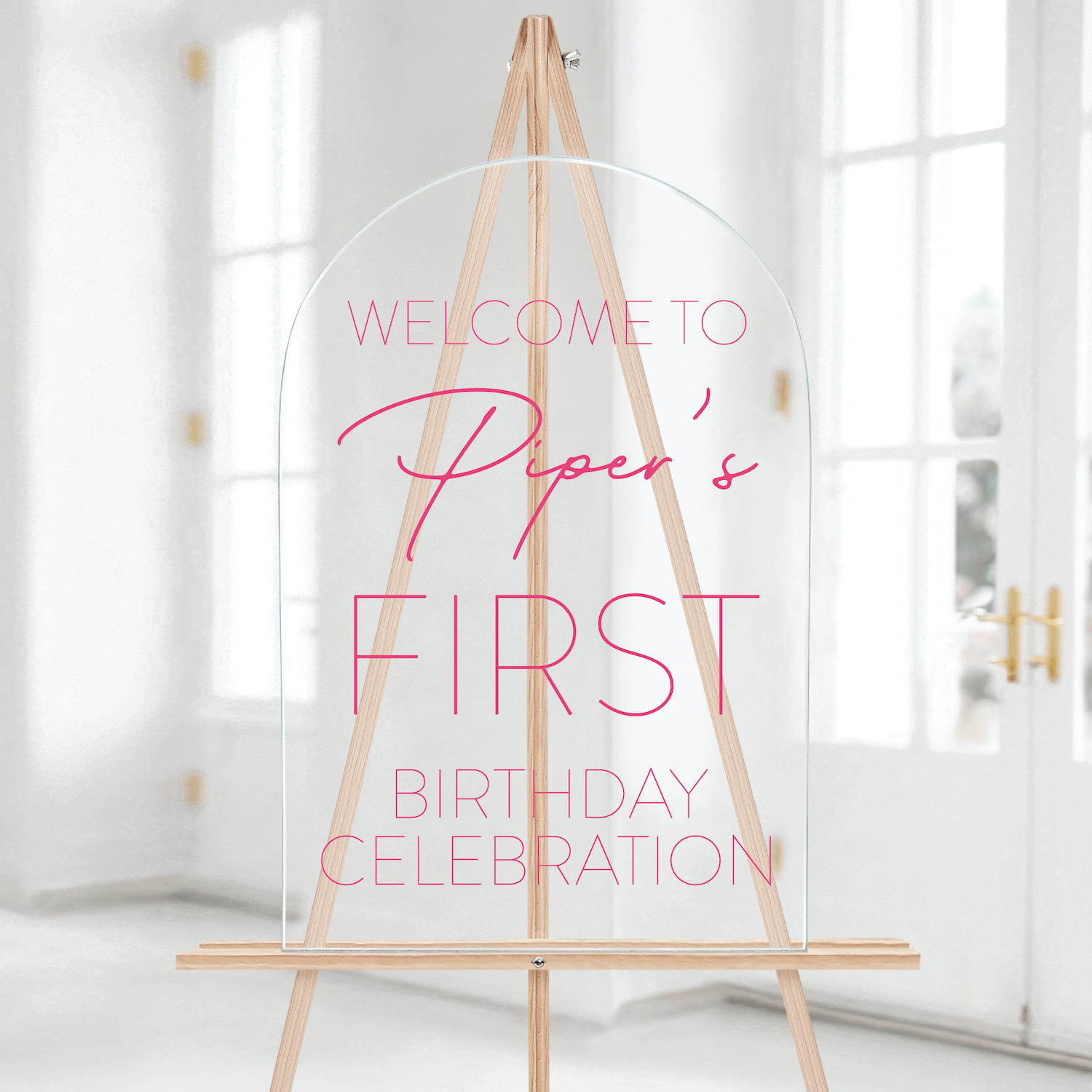 Wood easel with clear acrylic sign reading "Welcome to Piper's First Birthday Celebration" in pink text.