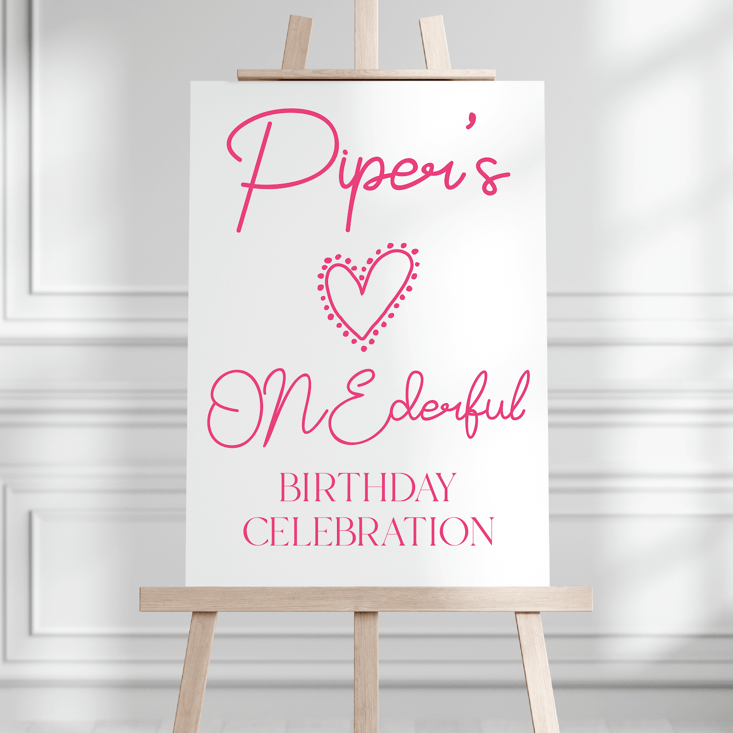 White welcome sign with pink script text reading "Piper's ONEderful BIRTHDAY CELEBRATION" and a dotted heart design, displayed on wooden easel.