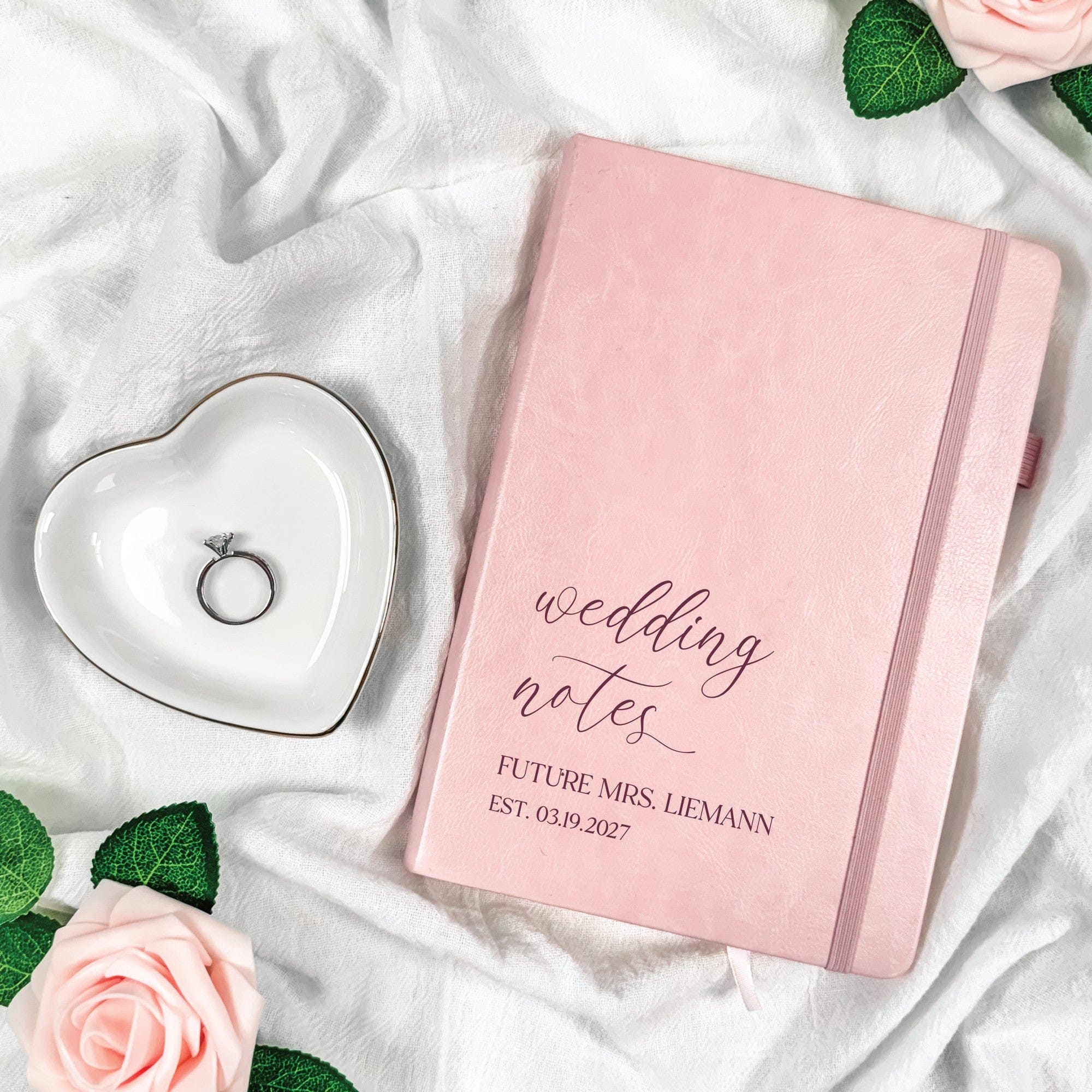 A pink personalized faux leather wedding notebook with an elastic band closure and a ribbon bookmark, displayed next to a heart-shaped ring dish on white fabric.