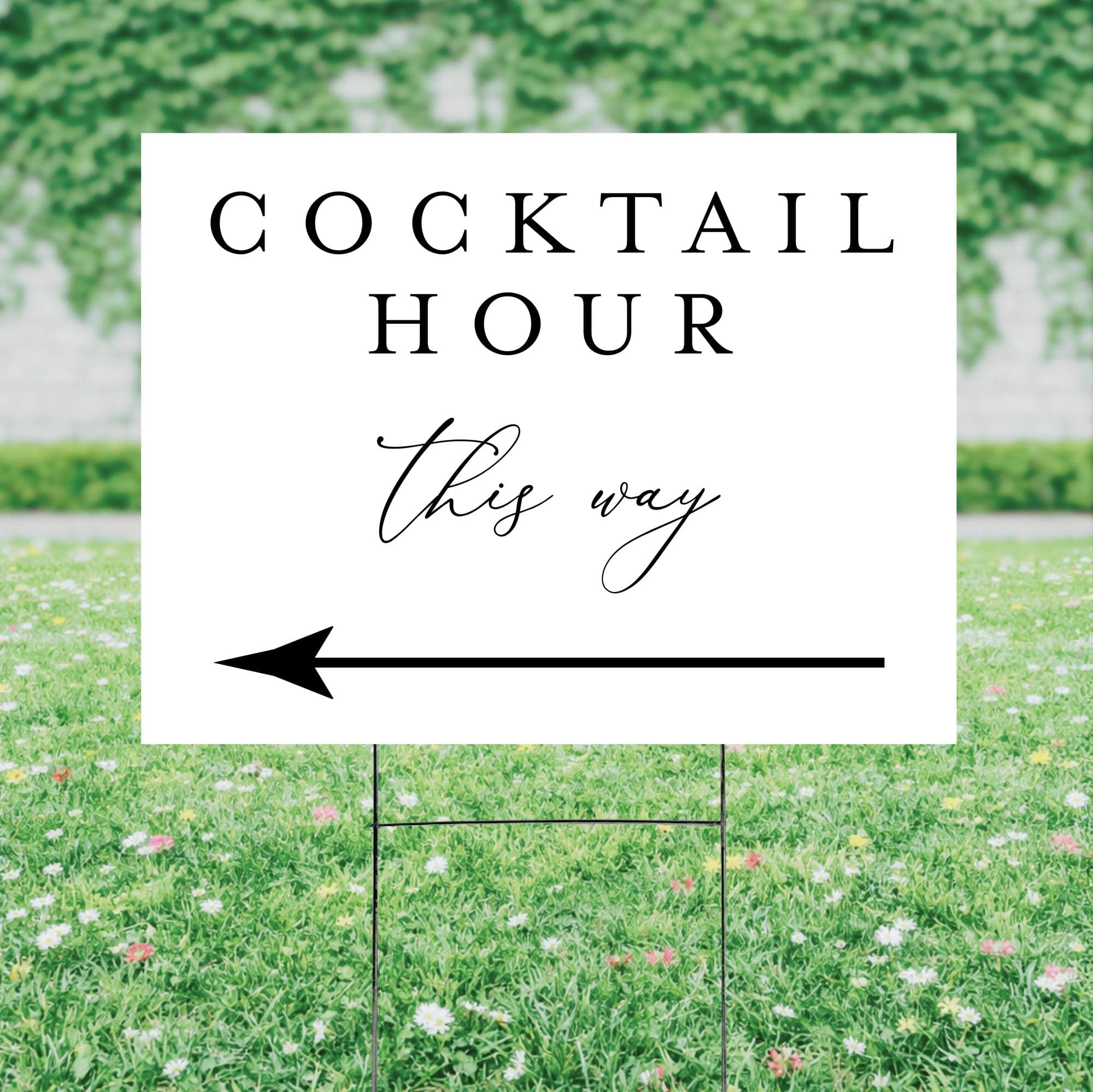 A white corrugated plastic yard sign on a metal H-stake, featuring black text and a left-pointing arrow, directs guests to a cocktail hour at an outdoor event.