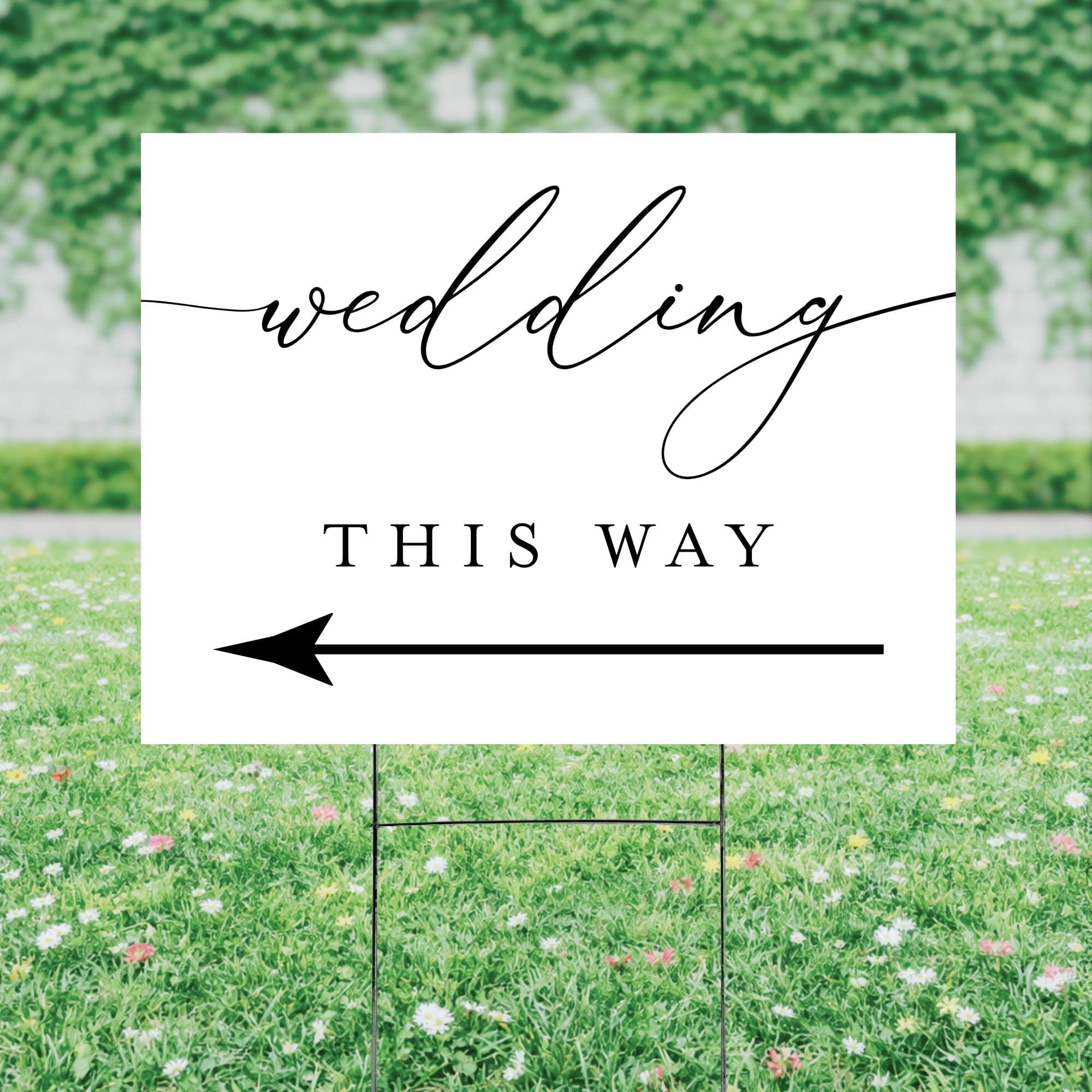 A rectangular white outdoor yard sign featuring a prominent black graphic and arrow, supported by a metal H-stake, serving to guide guests at a wedding celebration.