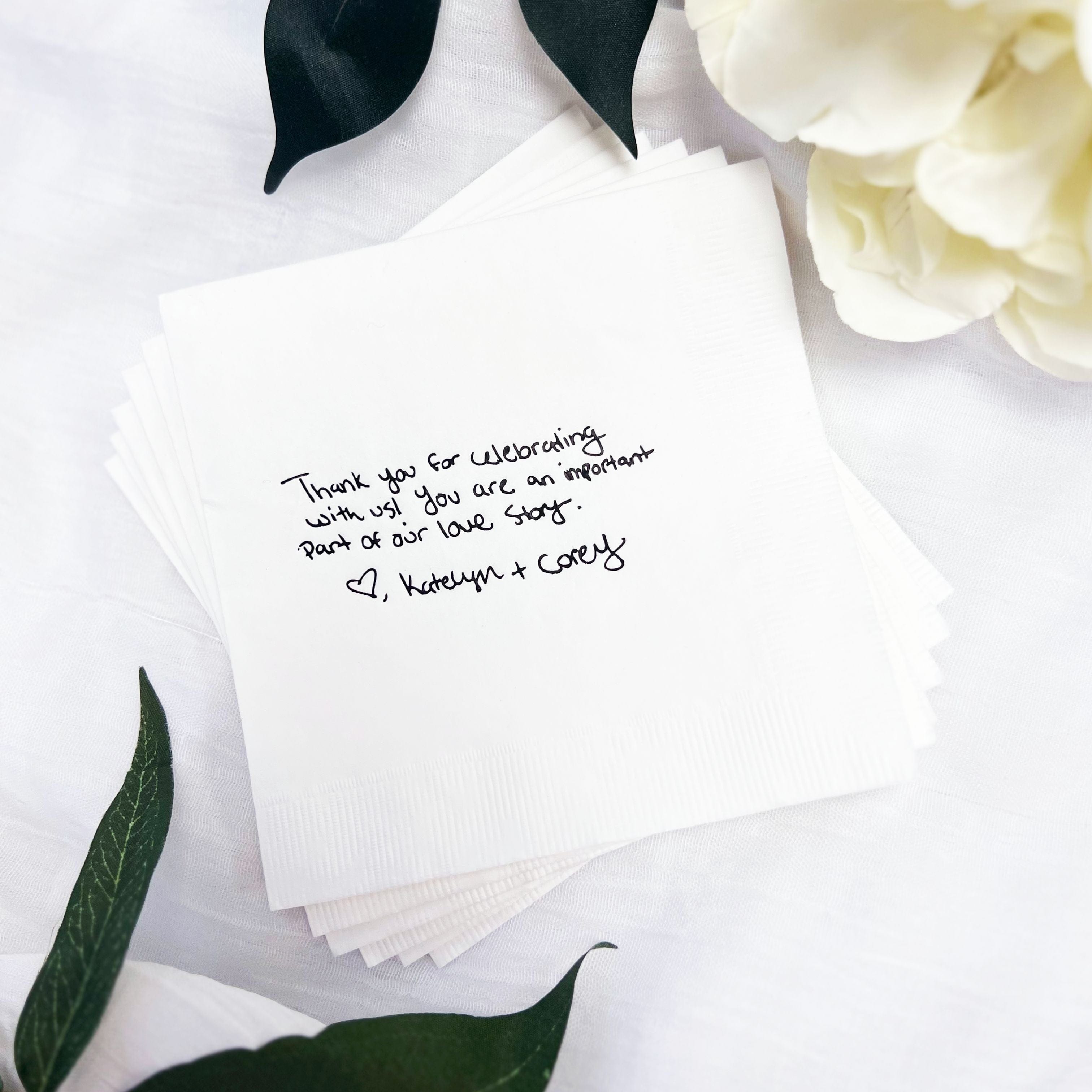Custom white paper wedding napkins featuring a personalized thank you message in a handwritten style, elegantly arranged with green foliage and a white flower.