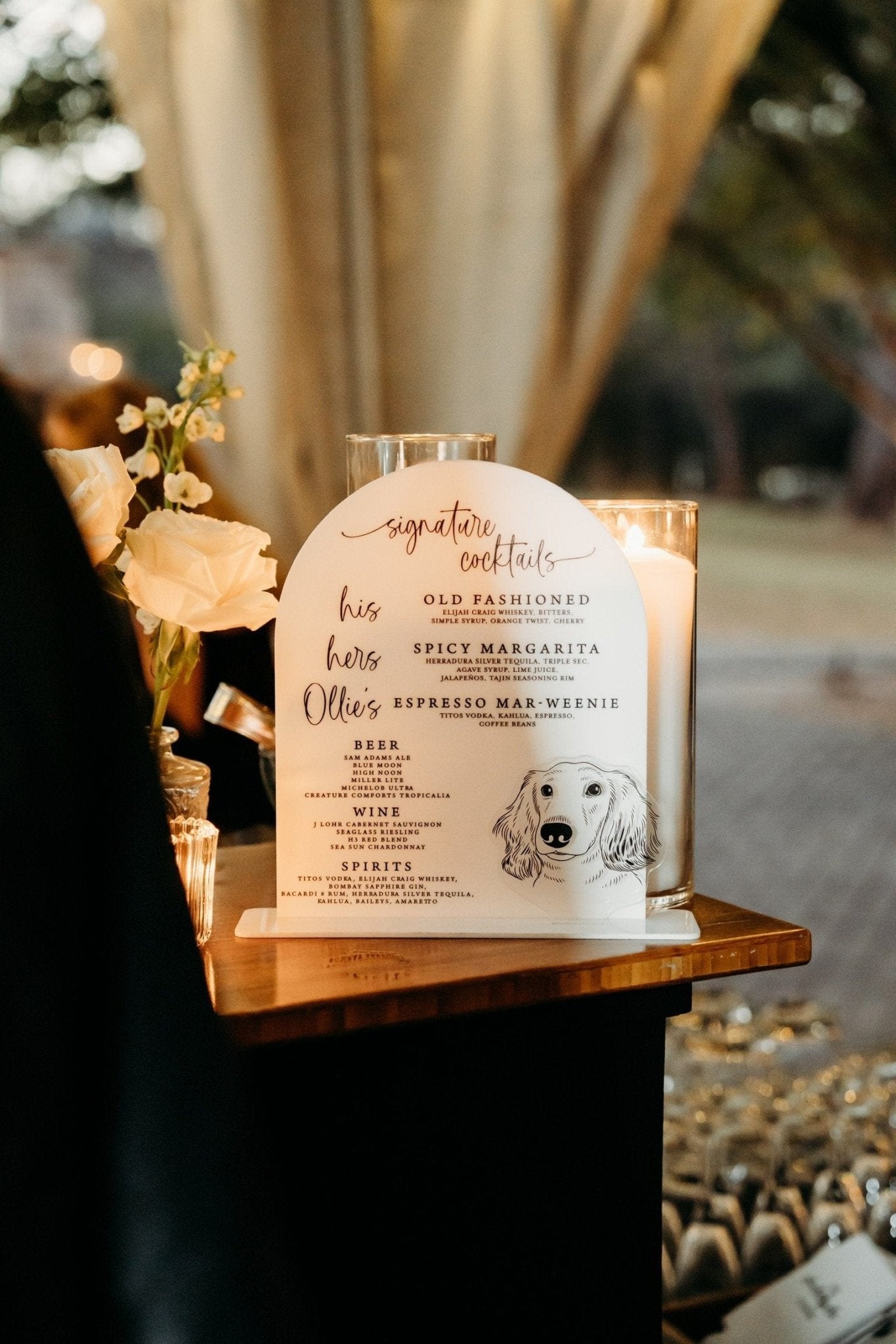An elegant white arched acrylic bar menu, showcasing a custom pet illustration and curated drink offerings, beautifully set amidst a refined celebration.