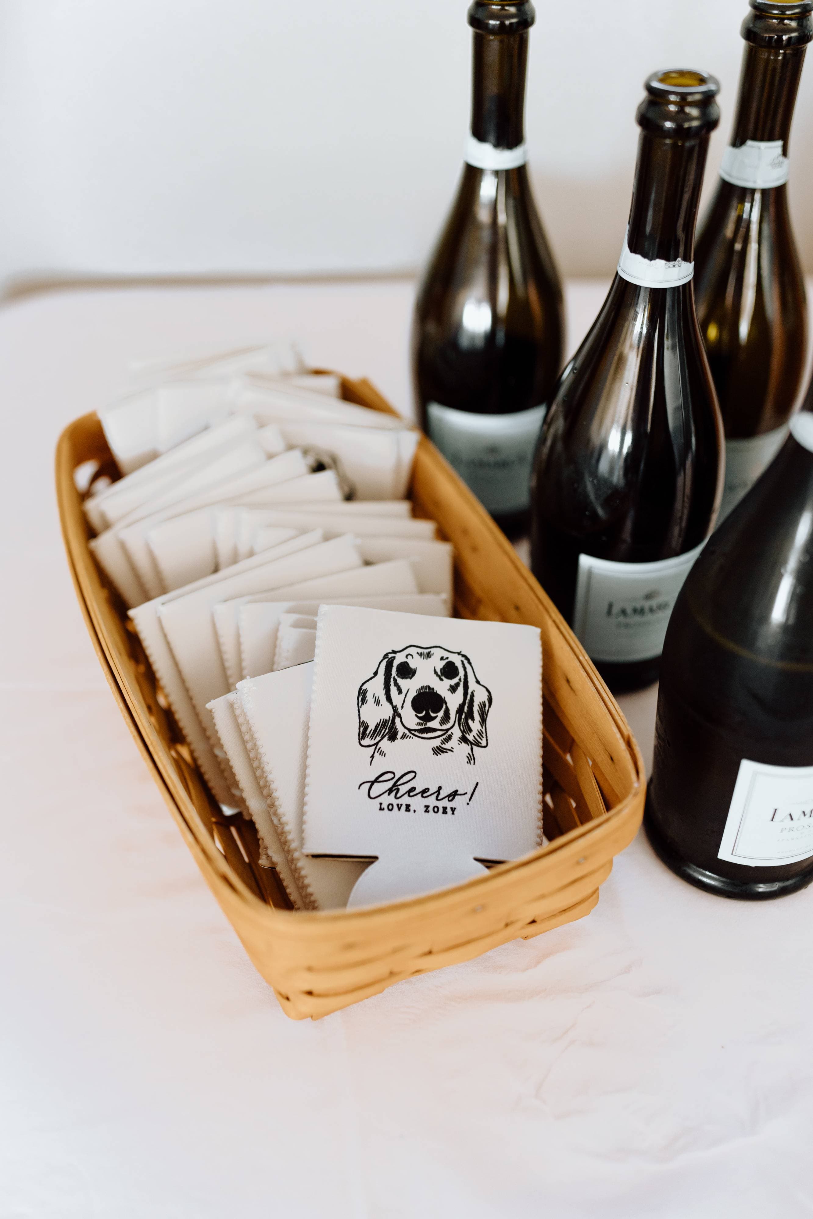 A woven basket holds white foam can coolers featuring a black dog illustration and a celebratory message, displayed alongside sparkling wine bottles as custom wedding favors.