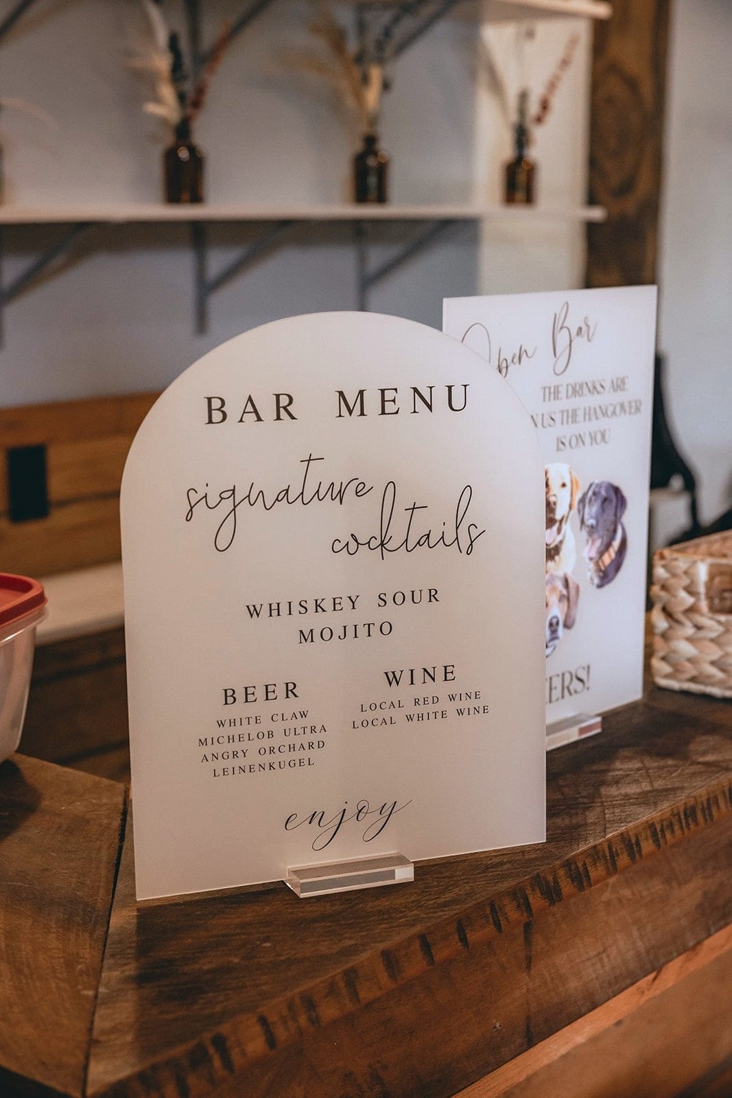 A modern frosted acrylic arched bar menu sign displaying signature cocktails, beer, and wine selections for a wedding or special event, placed on a rustic wooden bar.