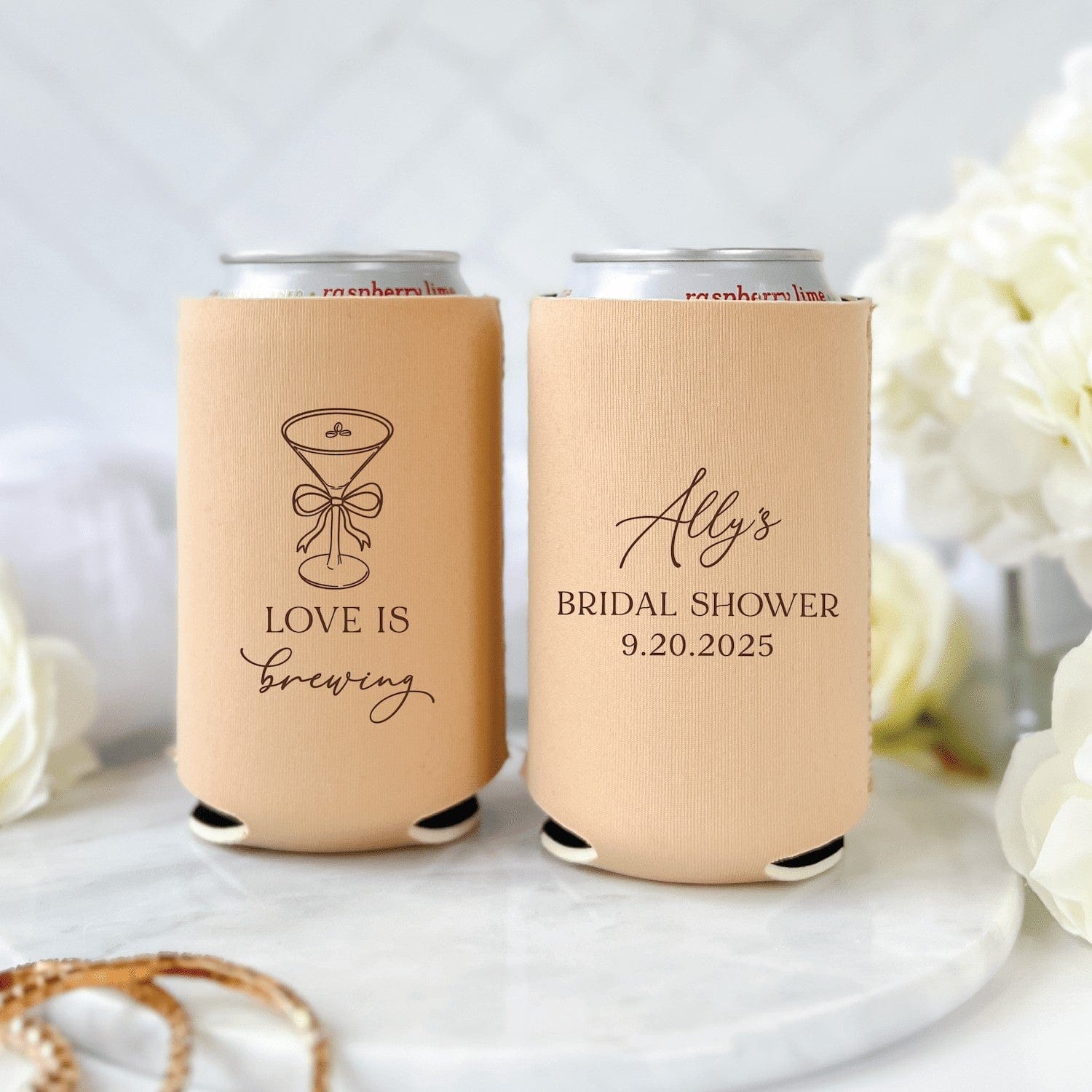 Two personalized peach neoprene can coolers for a bridal shower, one featuring a martini glass and love is brewing text, displayed on a marble surface with white flowers.