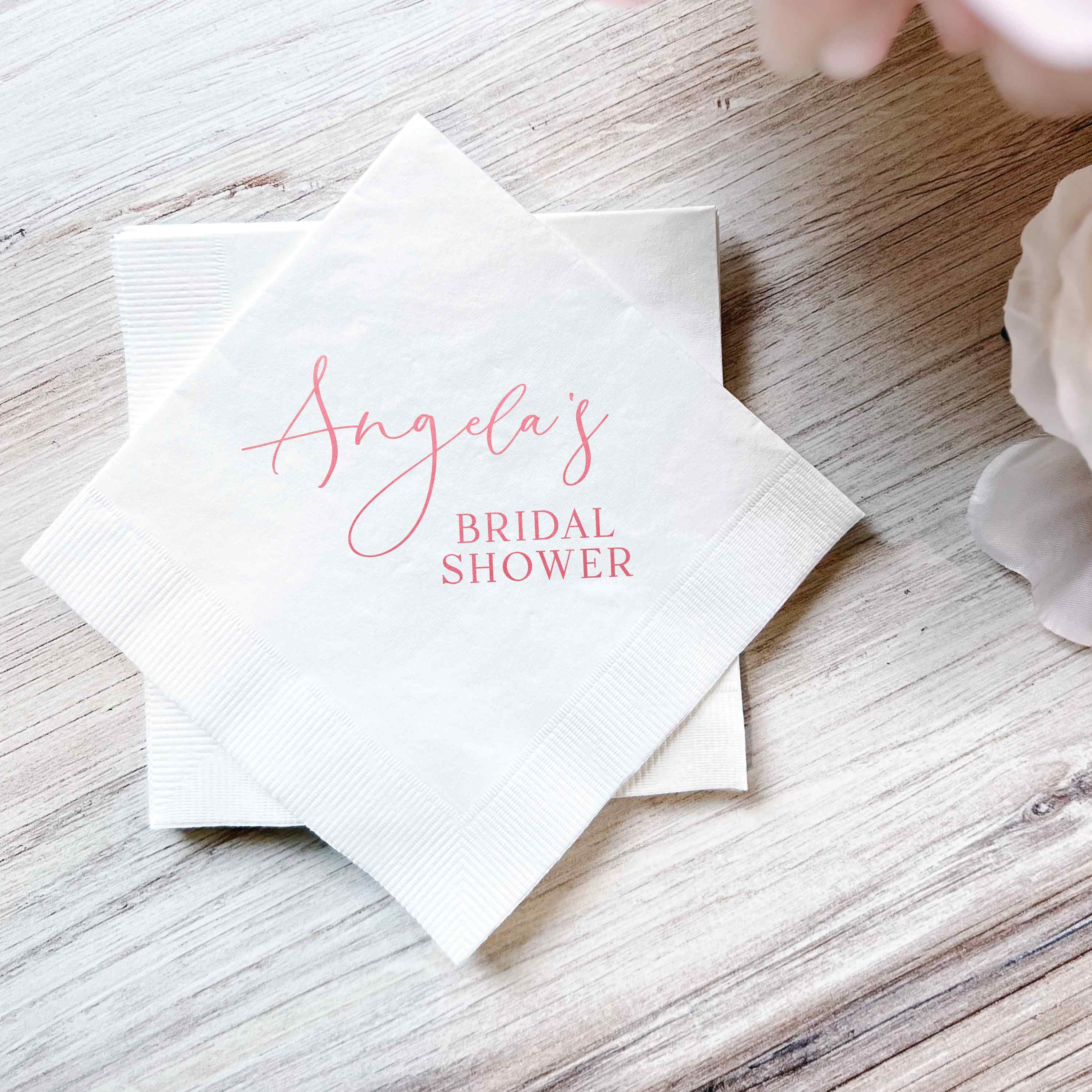 A stack of white paper bridal shower napkins personalized with pink script and block text, displayed on a light wooden surface.