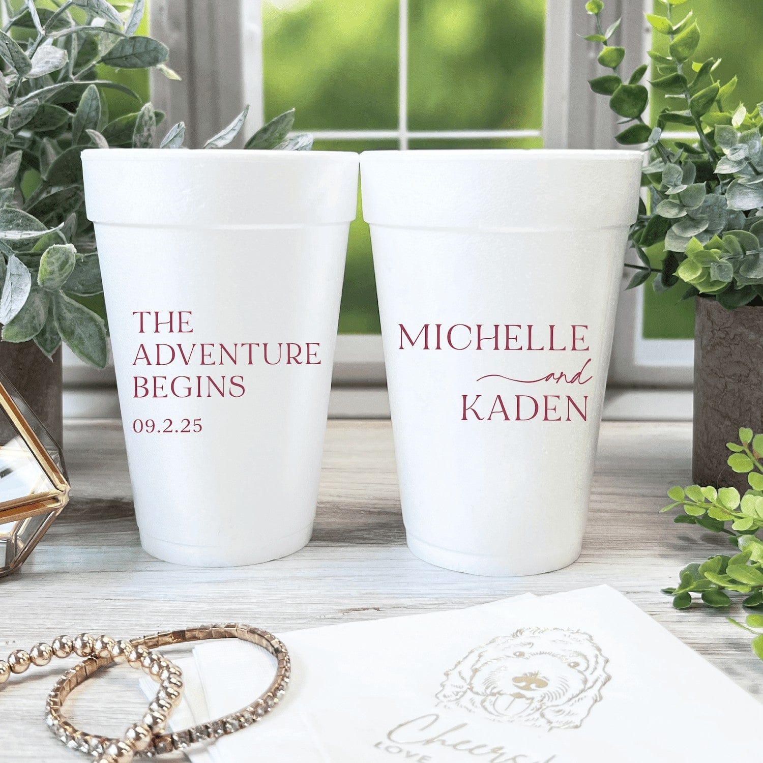 A pair of white personalized foam cups with custom wedding phrases, displayed on a rustic wooden table with decorative plants and jewelry.