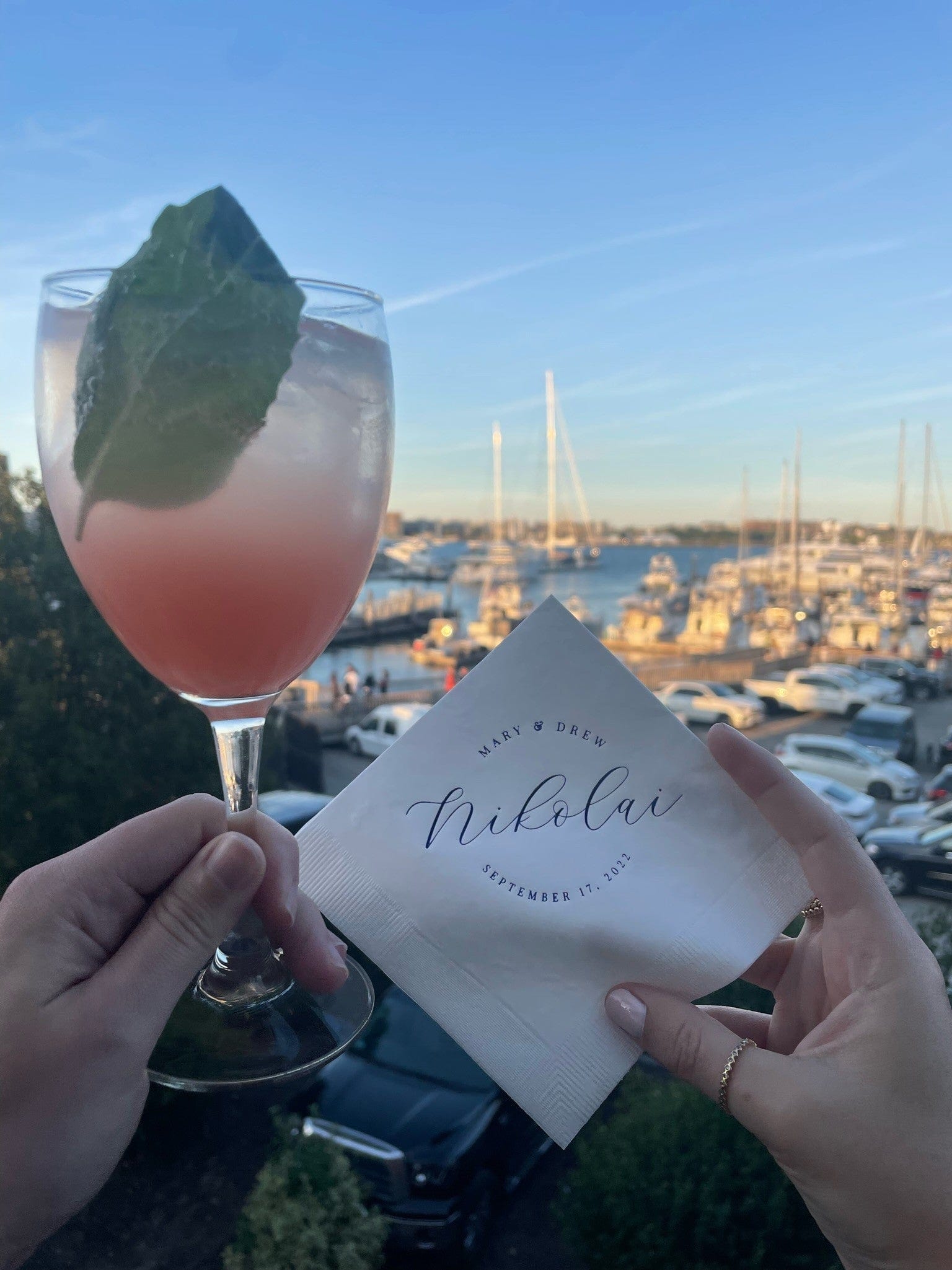 A person holds a pink cocktail and a white personalized paper napkin with custom script text at an outdoor wedding reception overlooking a marina with boats.
