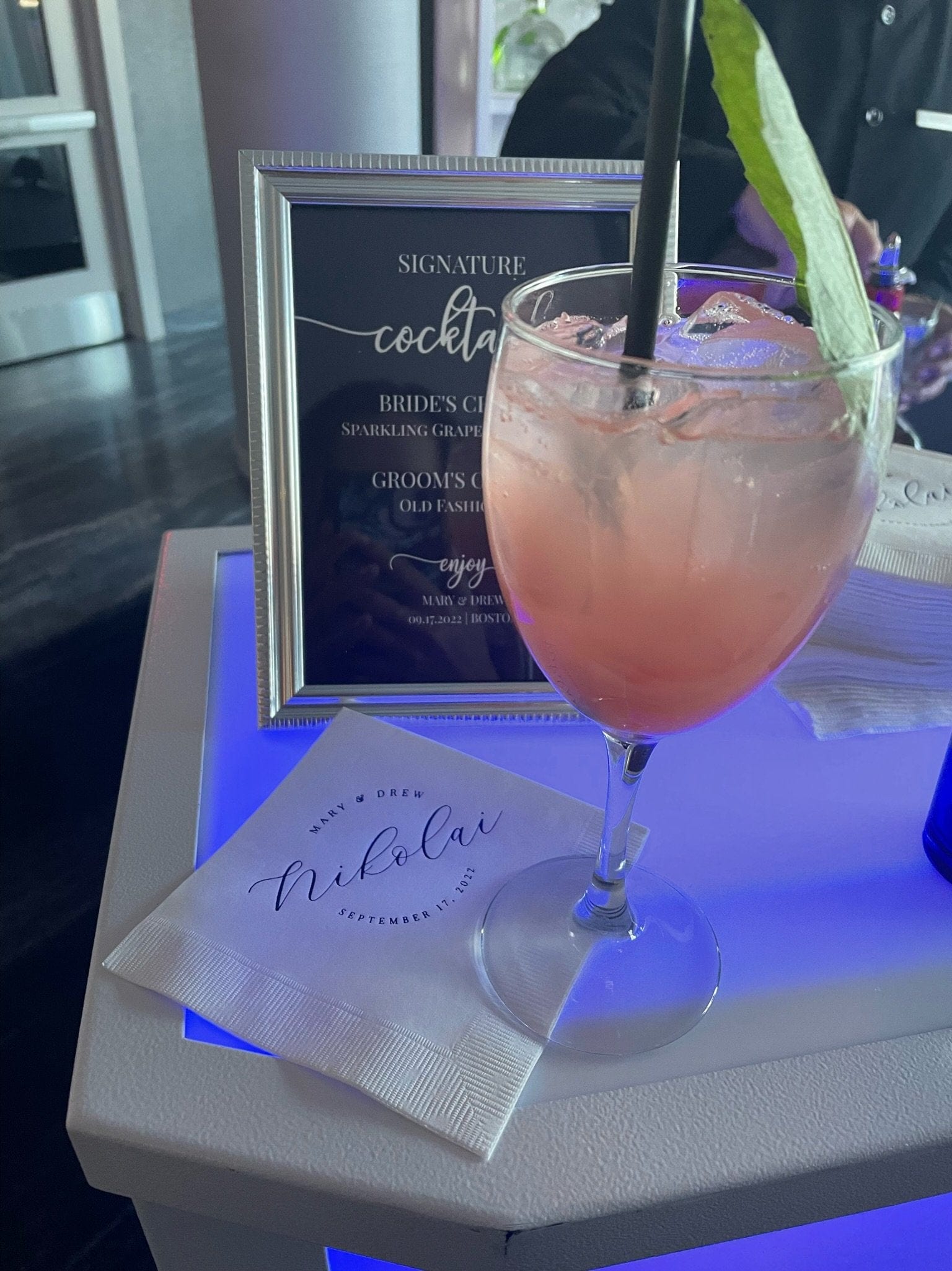 A personalized white paper wedding cocktail napkin with custom black script displayed next to a framed signature drink menu and a pink cocktail, set on a glow-lit bar.