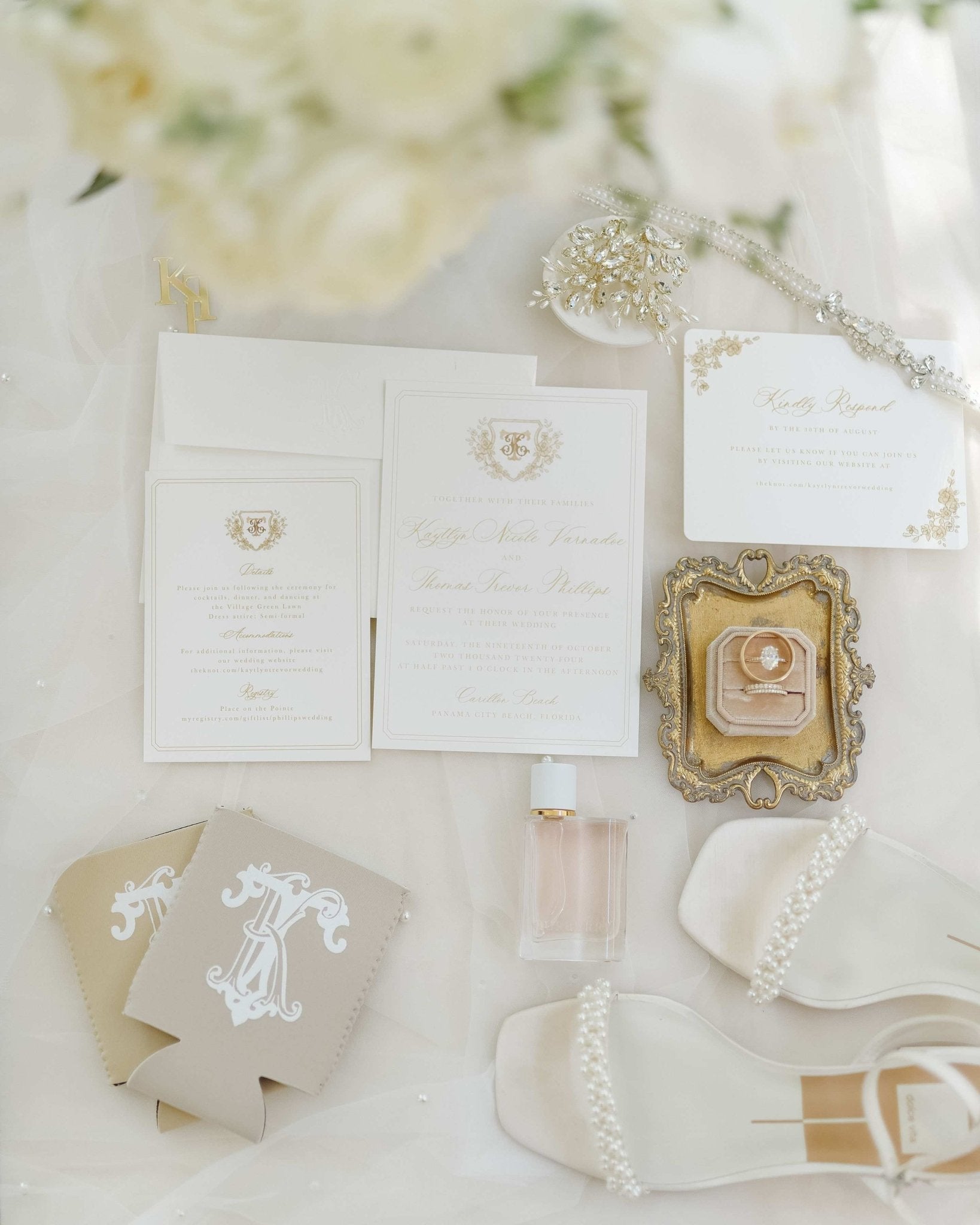 An elegant flat lay of cream and gold wedding stationery, engagement and wedding rings in a velvet box, white pearl-detailed bridal shoes, a perfume bottle, and custom tan beverage insulators.