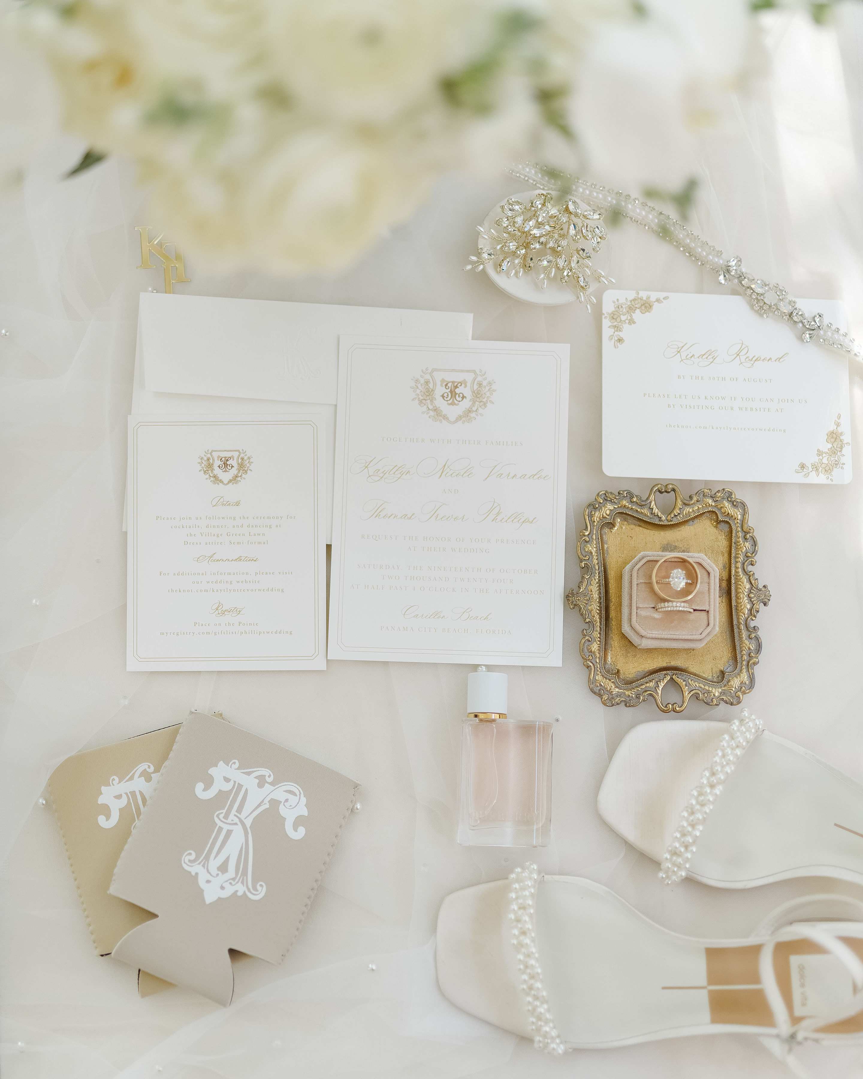 An elegant flat lay of cream and gold wedding stationery, engagement and wedding rings in a velvet box, white pearl-detailed bridal shoes, a perfume bottle, and custom tan beverage insulators.