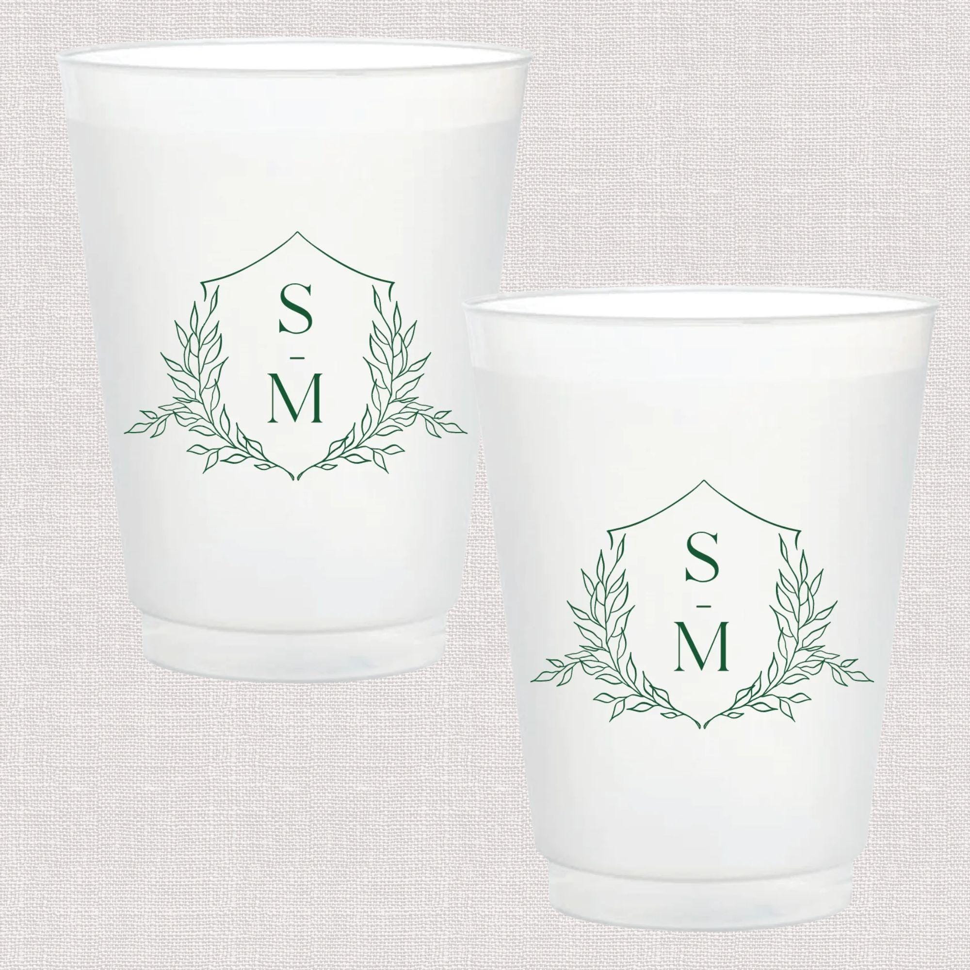 Two frosted white cups with sage green monogram design featuring initials S and M inside a diamond shield with laurel wreath accents.