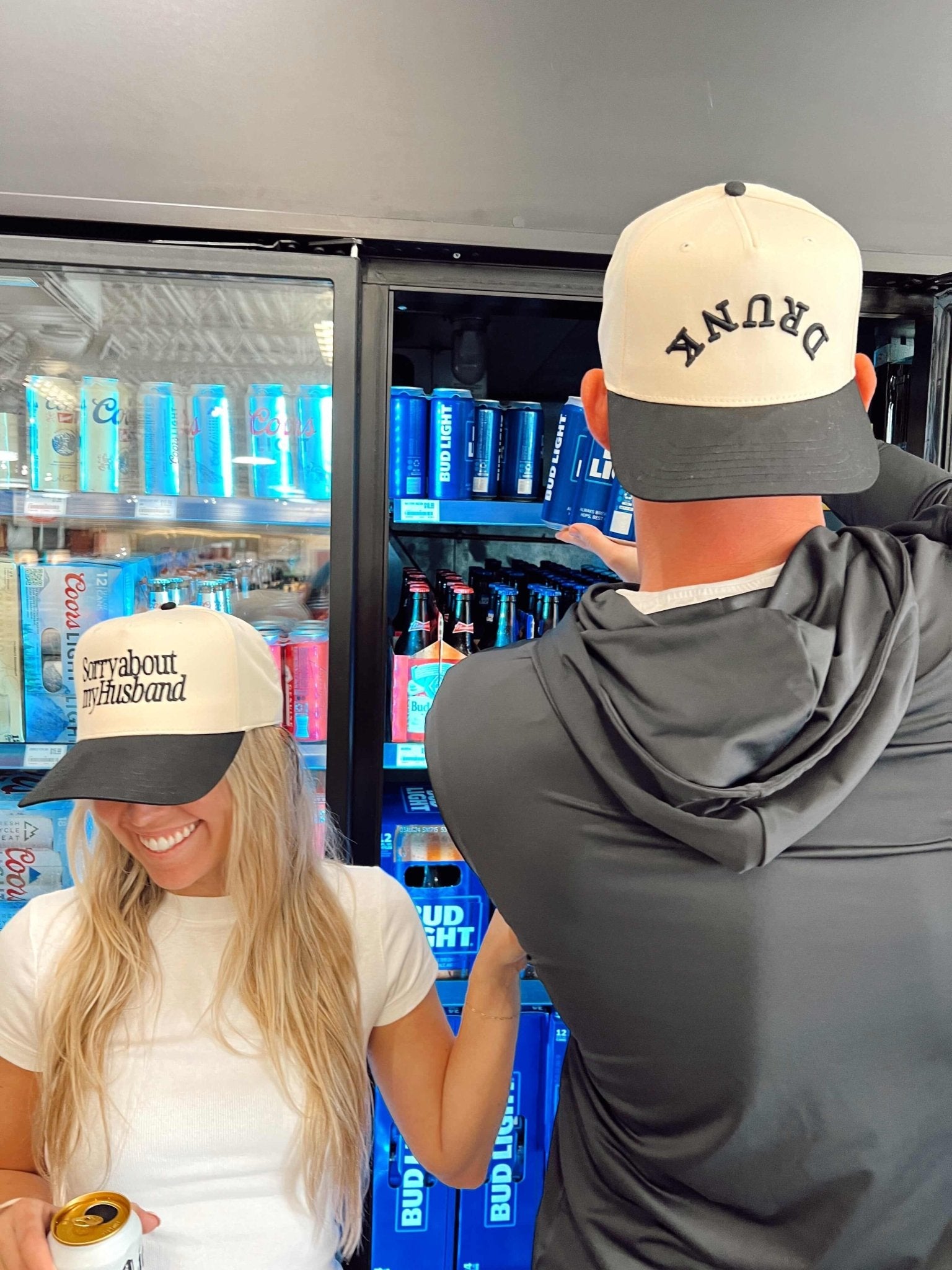 Two people wearing cream and charcoal vintage trucker hats with curved text logo design in front of a refrigerated beverage display.