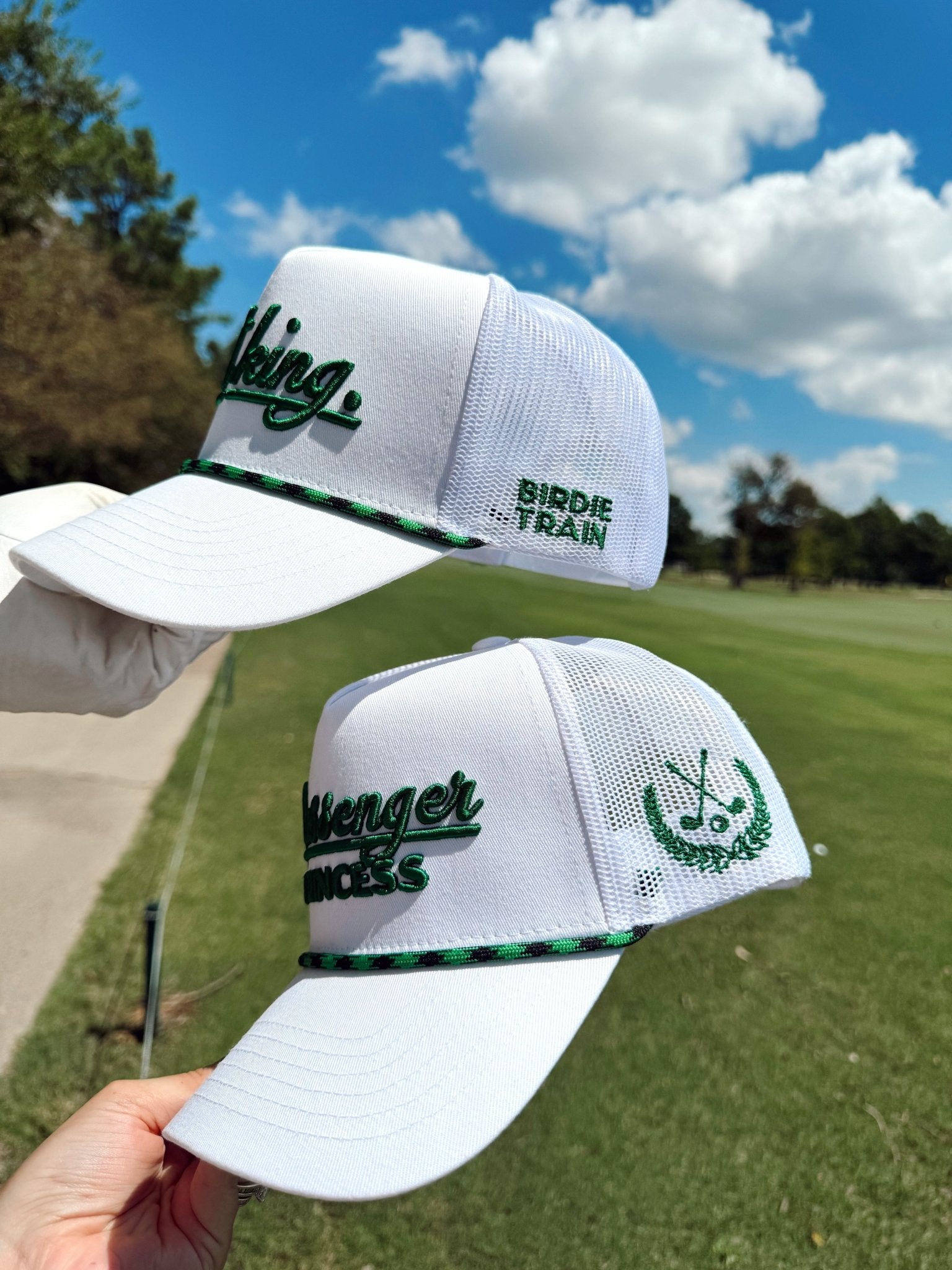 White mesh trucker hats with green embroidered golf-themed text and logos, displayed on a golf course background.