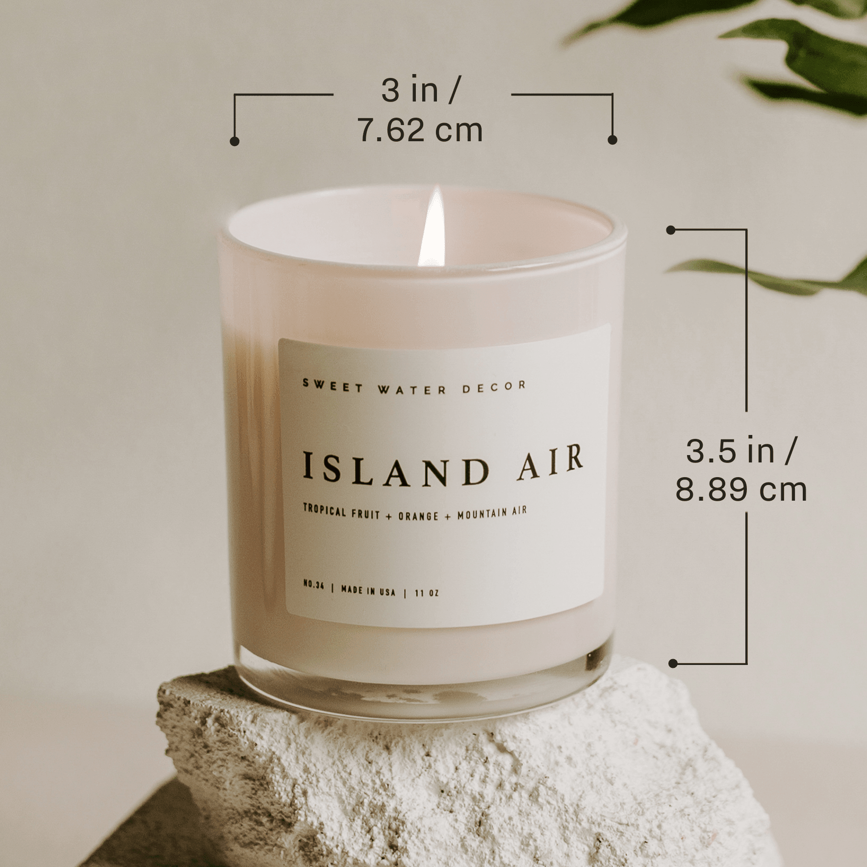 White soy candle labeled "Island Air" in glass jar with measurements shown, sitting on white stone with greenery in background.